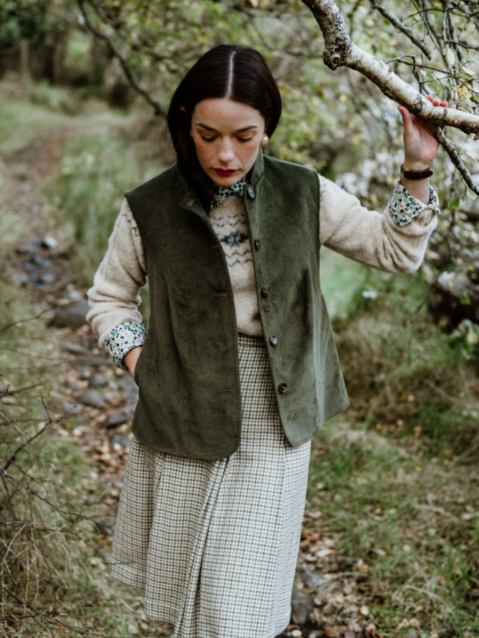 A dark-haired woman strolls outdoors in a Campbell's of Beauly Corduroy Gilet, paired with a patterned sweater and check skirt, holding a branch on a leafy path that radiates country elegance.