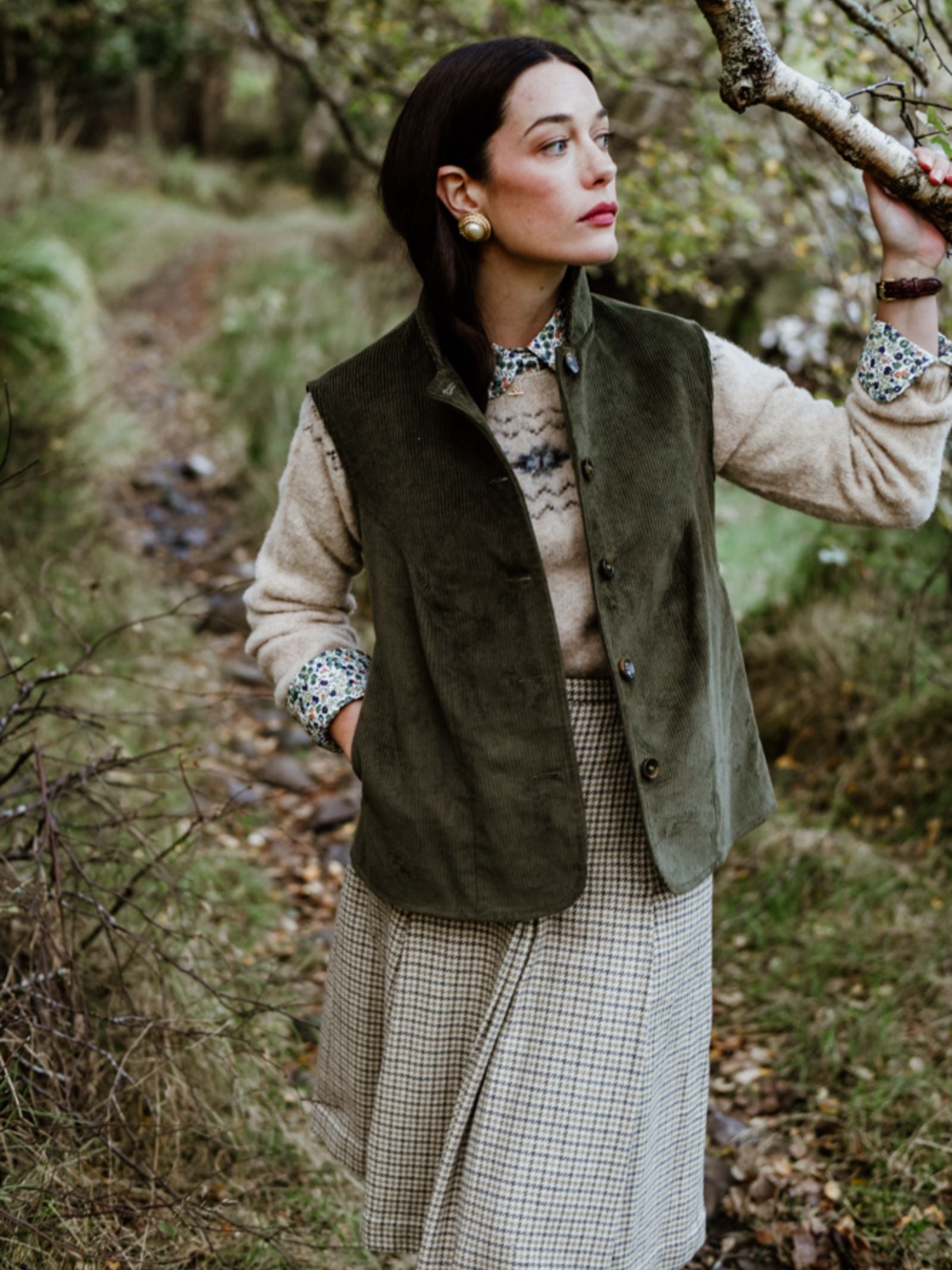 A dark-haired woman stands outdoors by a tree, exuding rural charm in the Corduroy Gilet by Campbell's of Beauly, layered over a patterned sweater, plaid skirt, and floral shirt, with green grass and a narrow path behind her.