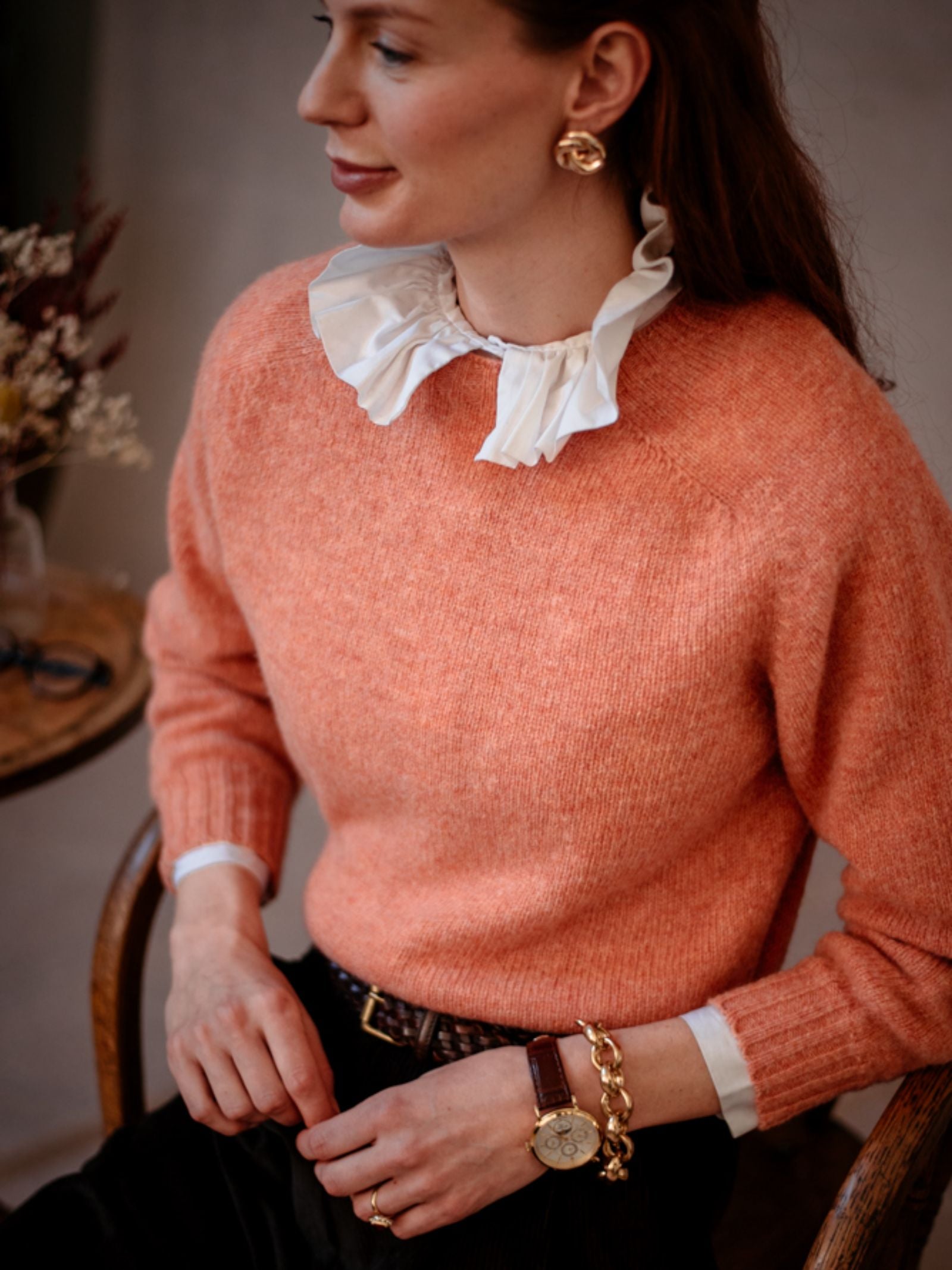 A woman sits on a chair wearing Campbells of Beaulys Cropped Shetland Jumper in peach with a ruffled white collar, gold earrings, bracelet, and watch; dried flowers in the background highlight her timeless knit style.