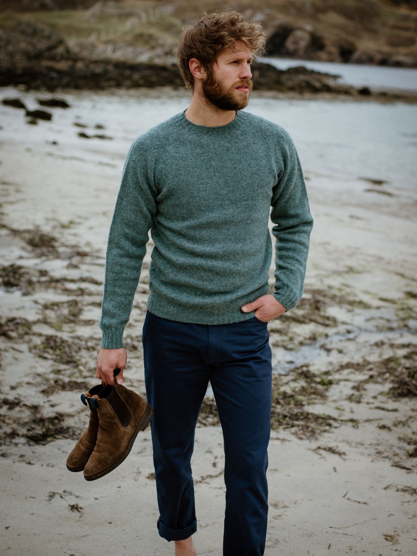 A bearded man stands barefoot on the beach, gazing sideways while holding brown suede boots. He wears the Campbells of Beauly Shetland Jumper and blue pants, with rocky hills and water completing the scenic backdrop.