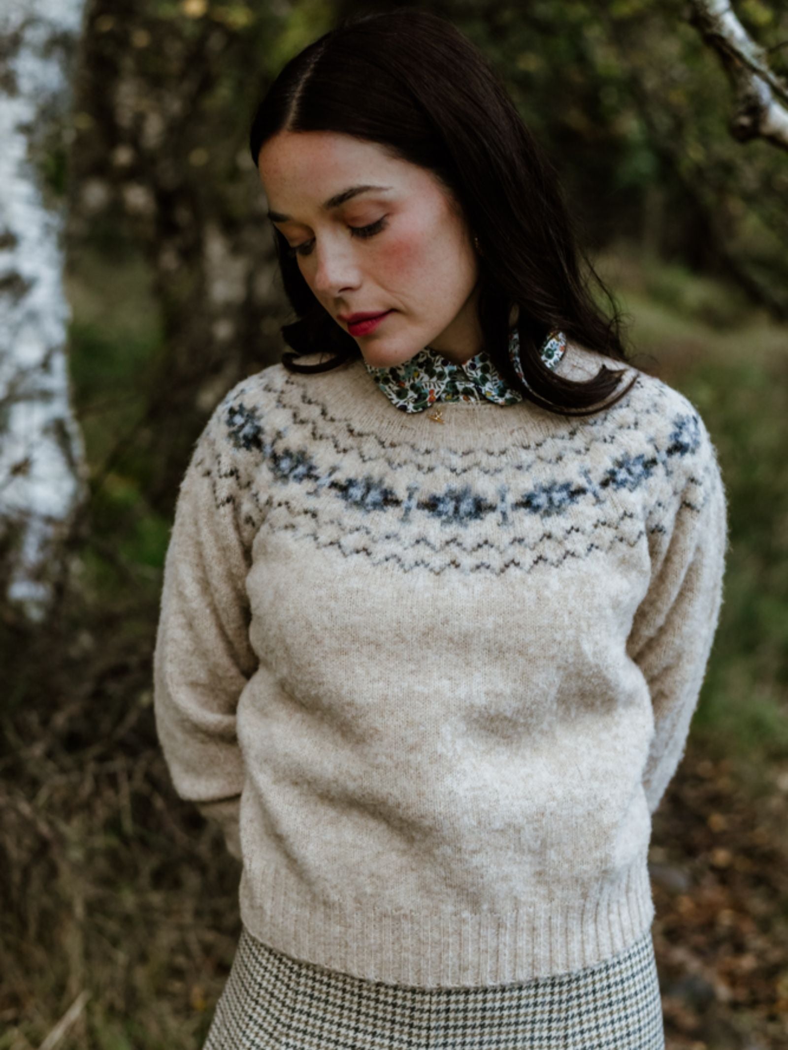 A woman with long dark hair stands outdoors among blurred greenery, wearing the Cropped Fairisle Crew Jumper by Campbell's of Beauly over a floral collared shirt and a houndstooth skirt.