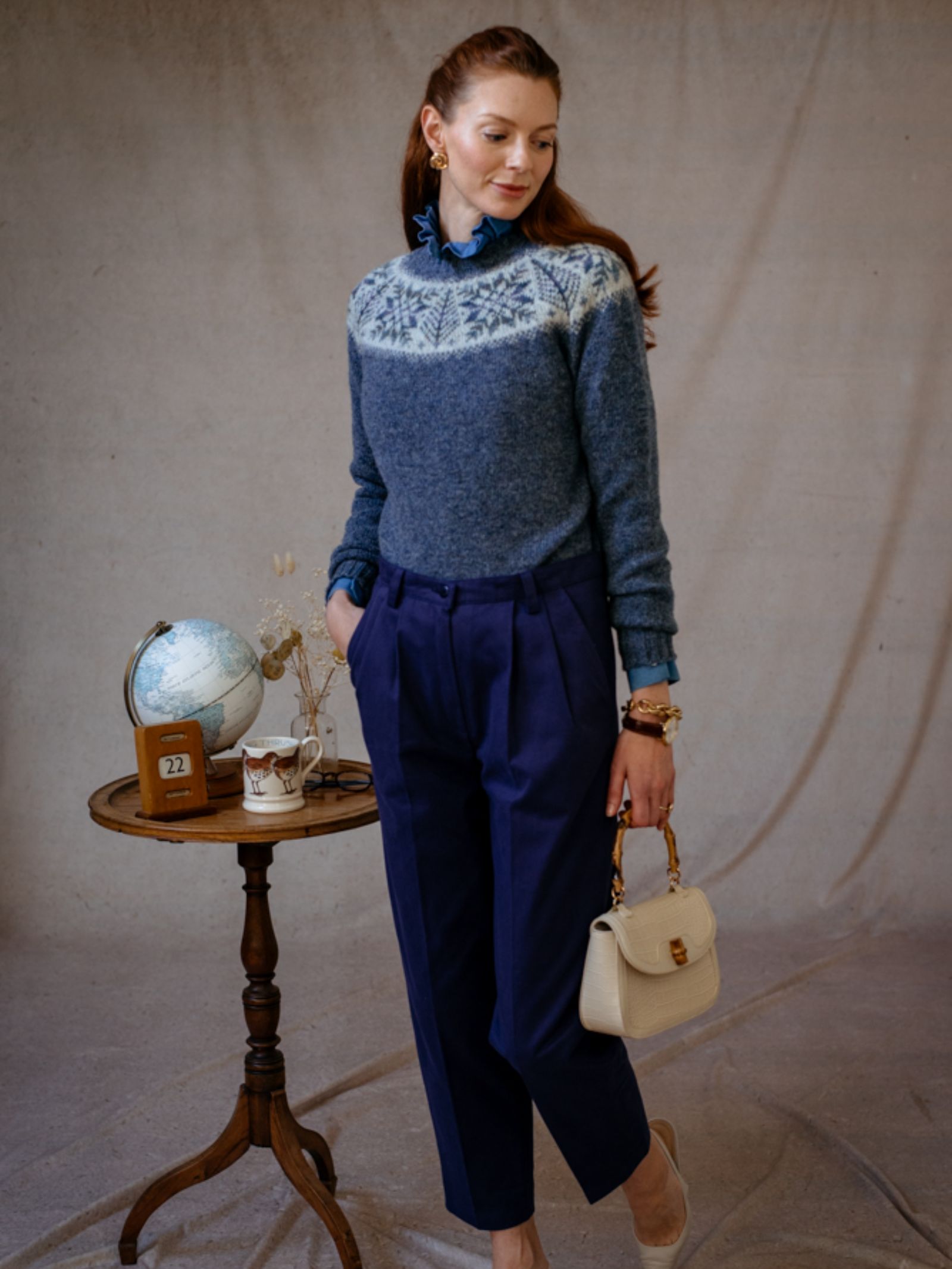A woman indoors stands by a round wooden table, smiling in a blue Shetland Wool Snowflake Crew Jumper by Campbell’s of Beauly, blue pleated trousers, and nude heels. She holds a cream handbag; a globe and calendar sit on the table.