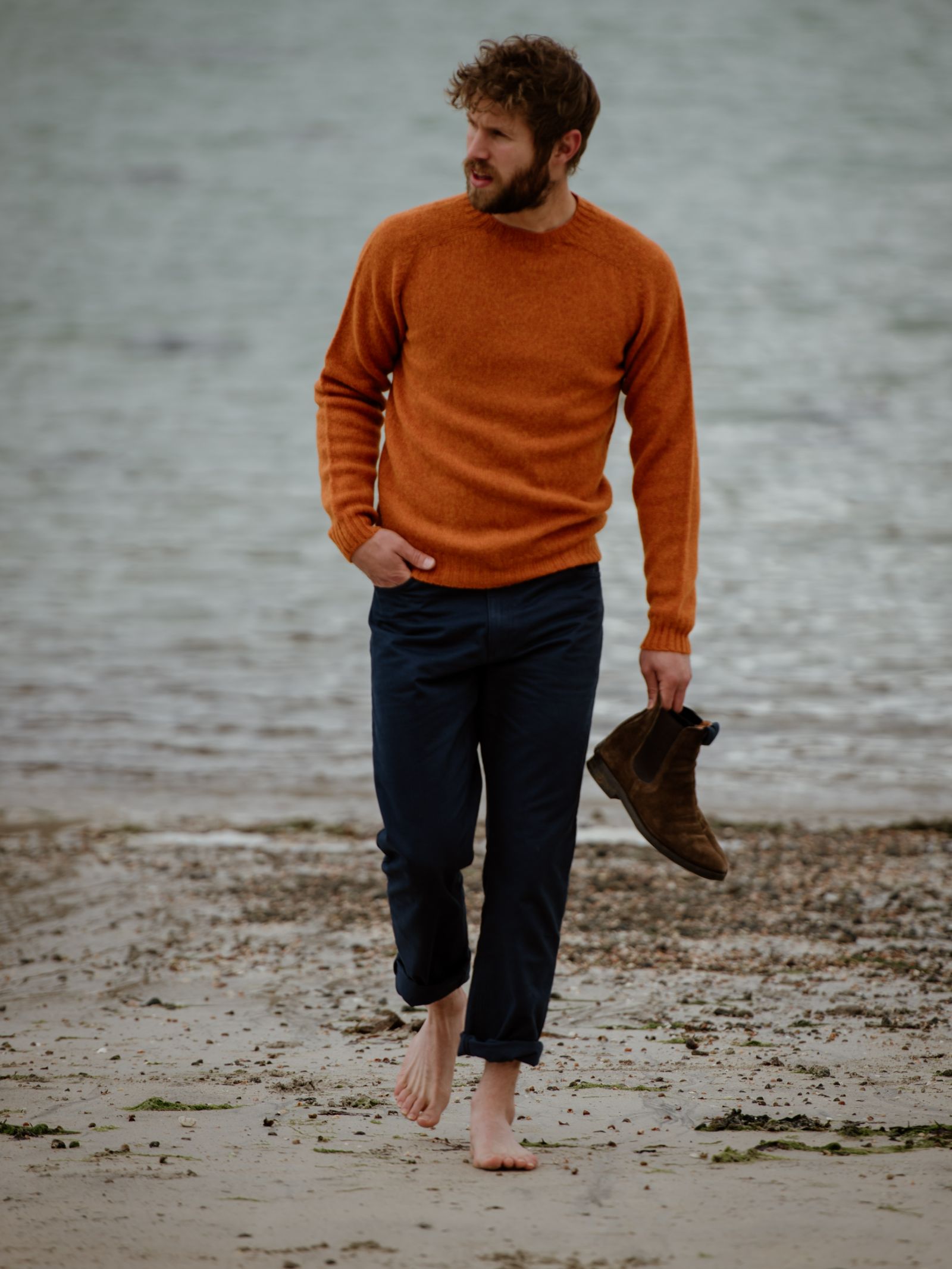 A bearded man, barefoot on the beach, holds brown boots and looks off to the side by the water. He wears dark pants and a Campbells of Beauly Shetland Jumper in heritage-inspired colourways.