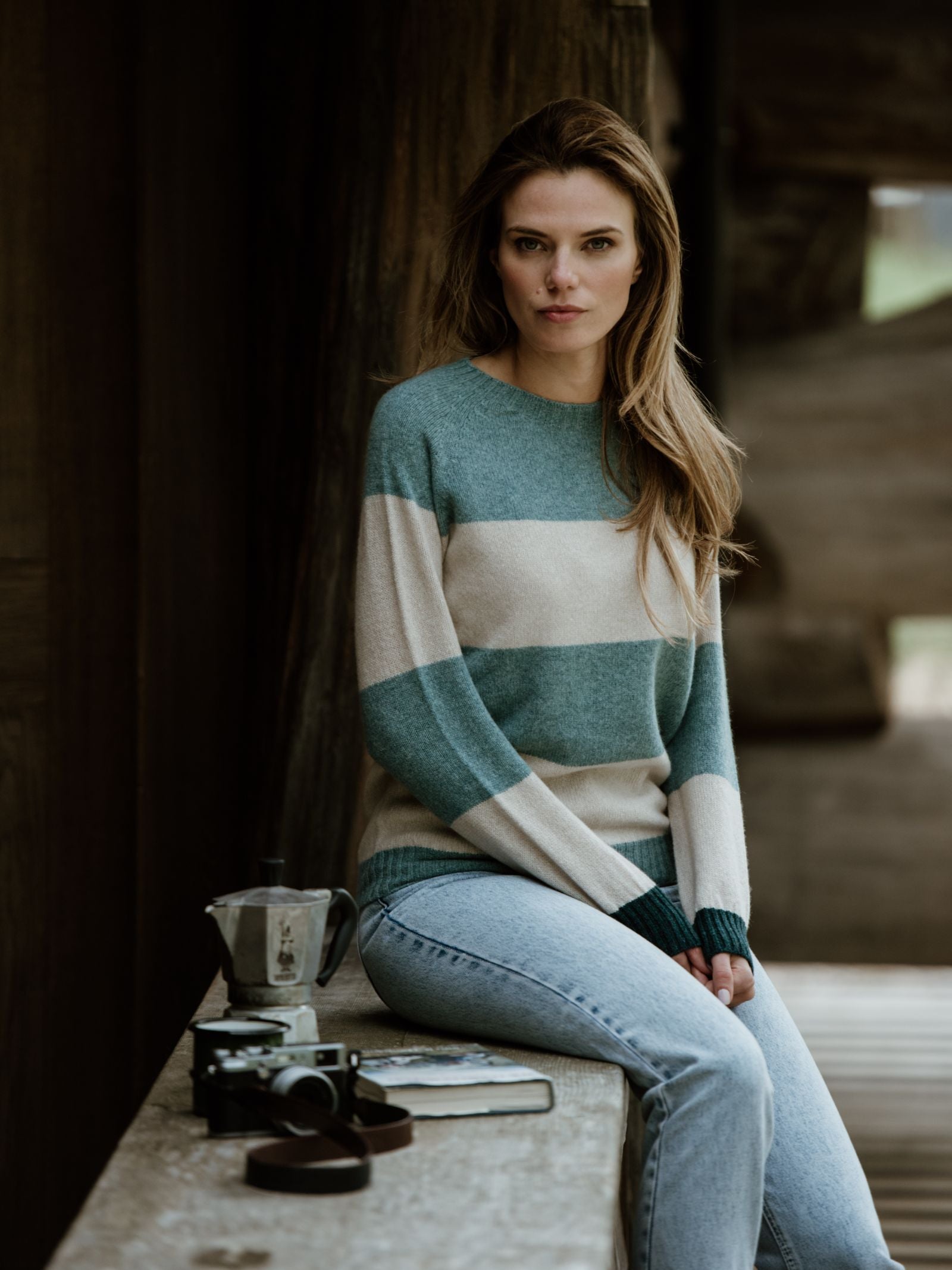 A woman in the Campbells of Beauly Stripe Crew Jumper sits on a wooden bench. The Scottish-made green and beige knit is paired with a camera, coffee maker, leather strap, and notebook in a rustic setting.