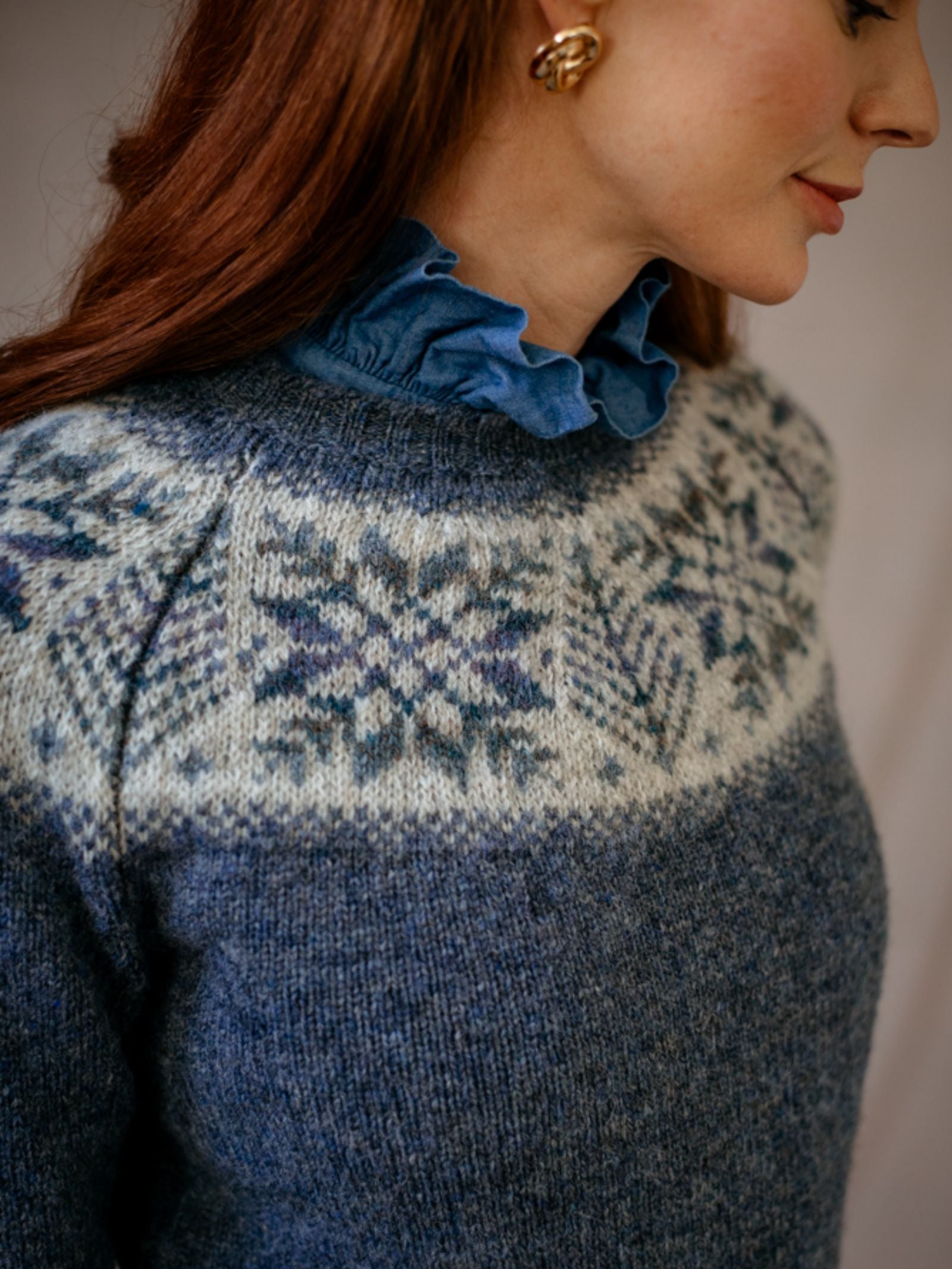 A woman with long red hair wears the Campbells of Beauly Shetland Wool Snowflake Crew Jumper over a blue ruffled collar blouse. She has gold earrings and is shown from the shoulders up, facing slightly right.