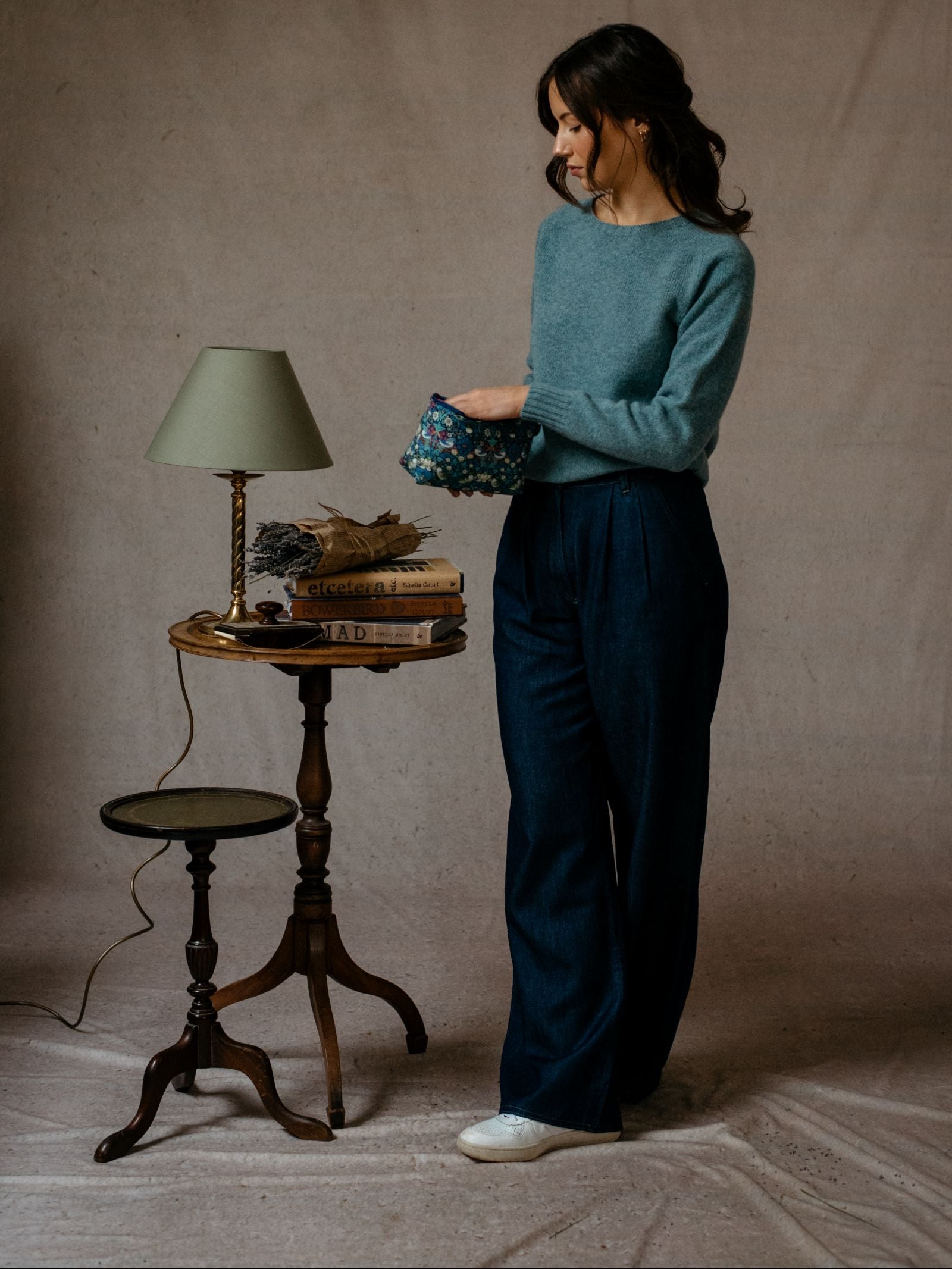 A woman wears a blue Campbells of Beauly Geelong Lambswool Crew Neck Jumper and wide-leg jeans, standing by a small table with books, dried flowers, and a lamp. She holds a floral pouch against a neutral backdrop.