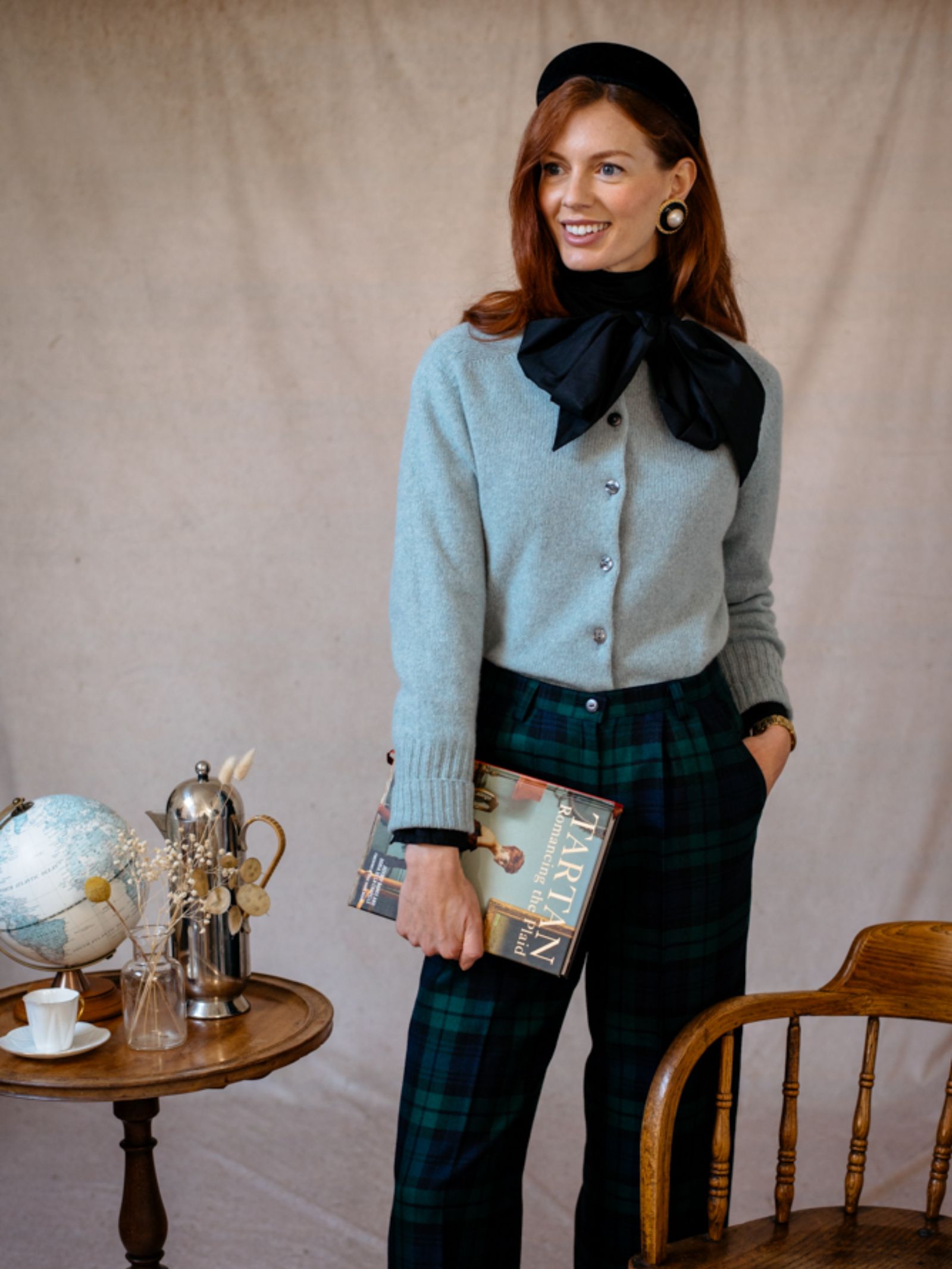 A woman with long red hair, in a gray cardigan, large black bow, Campbells of Beauly Wide Leg Blackwatch Trousers, and a black beret smiles while holding a book beside a small table with a globe, teapot, and teacup.