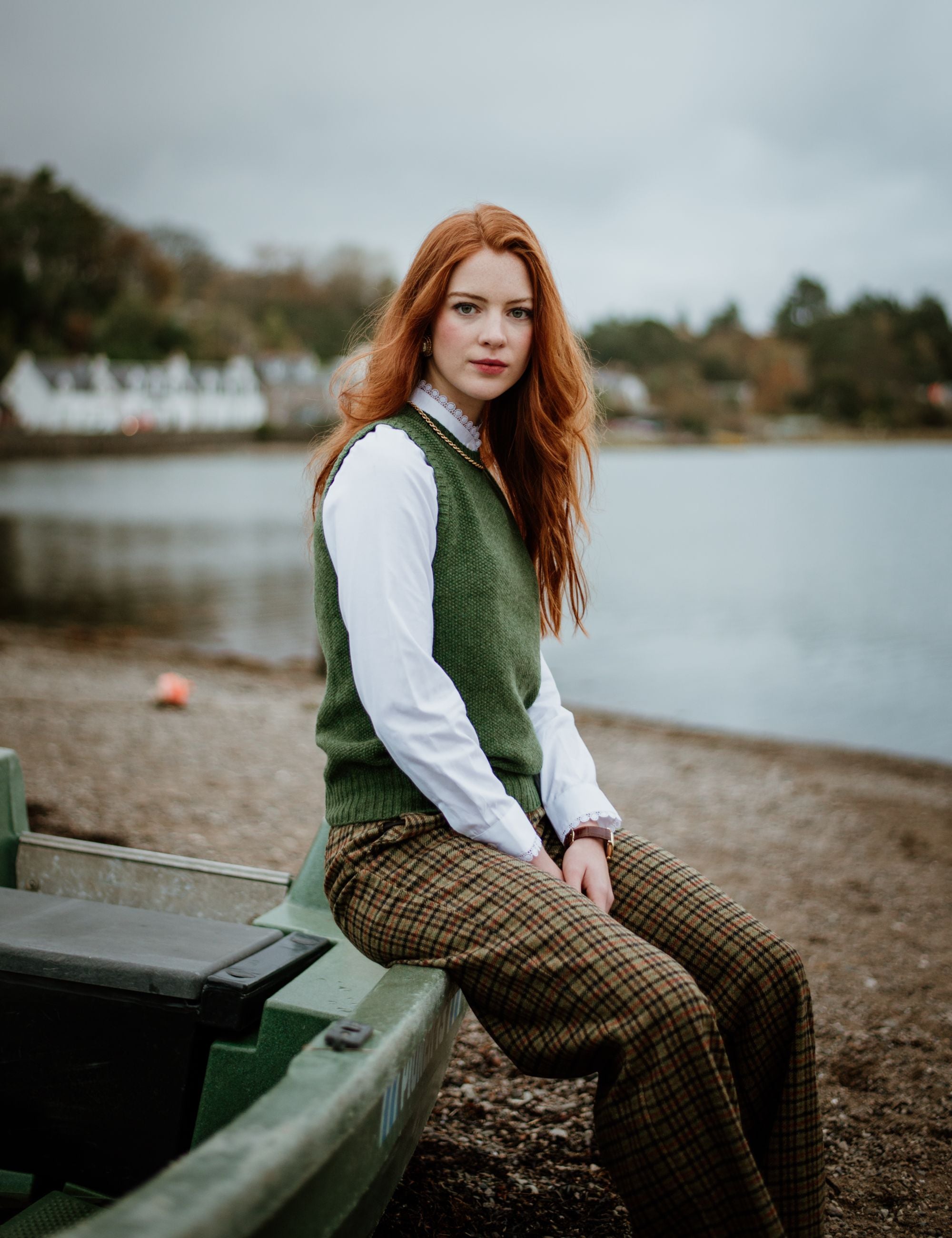 A woman with long red hair sits on the edge of a green rowboat by a lake, wearing a white shirt, green vest, and brown plaid pants. The background shows water, trees, and houses under a cloudy sky.