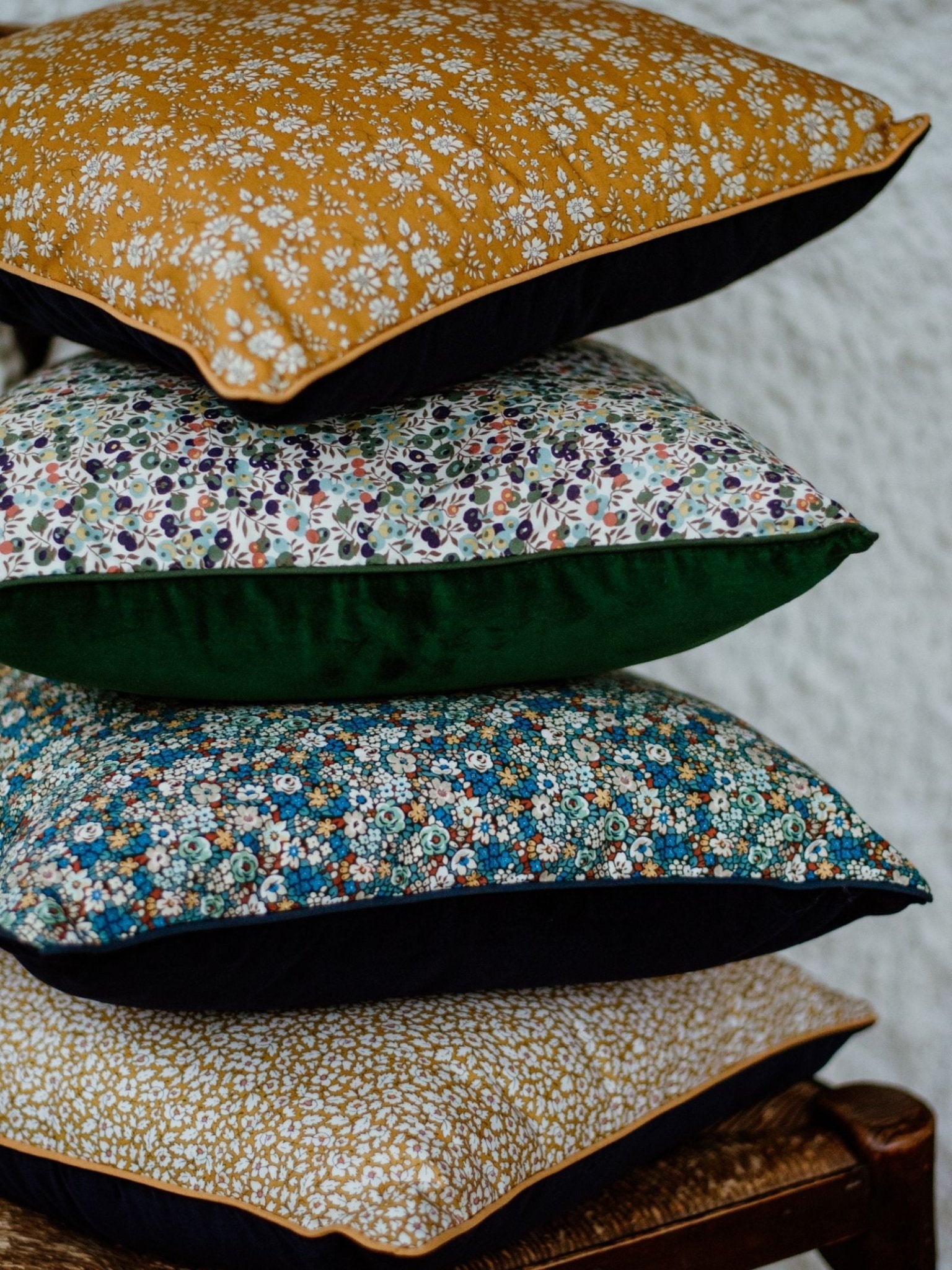 A stack of four Campbells of Beauly Liberty Print Cushion Covers in various yellow, green, and blue floral patterns rests on a wooden chair against a textured white wall.