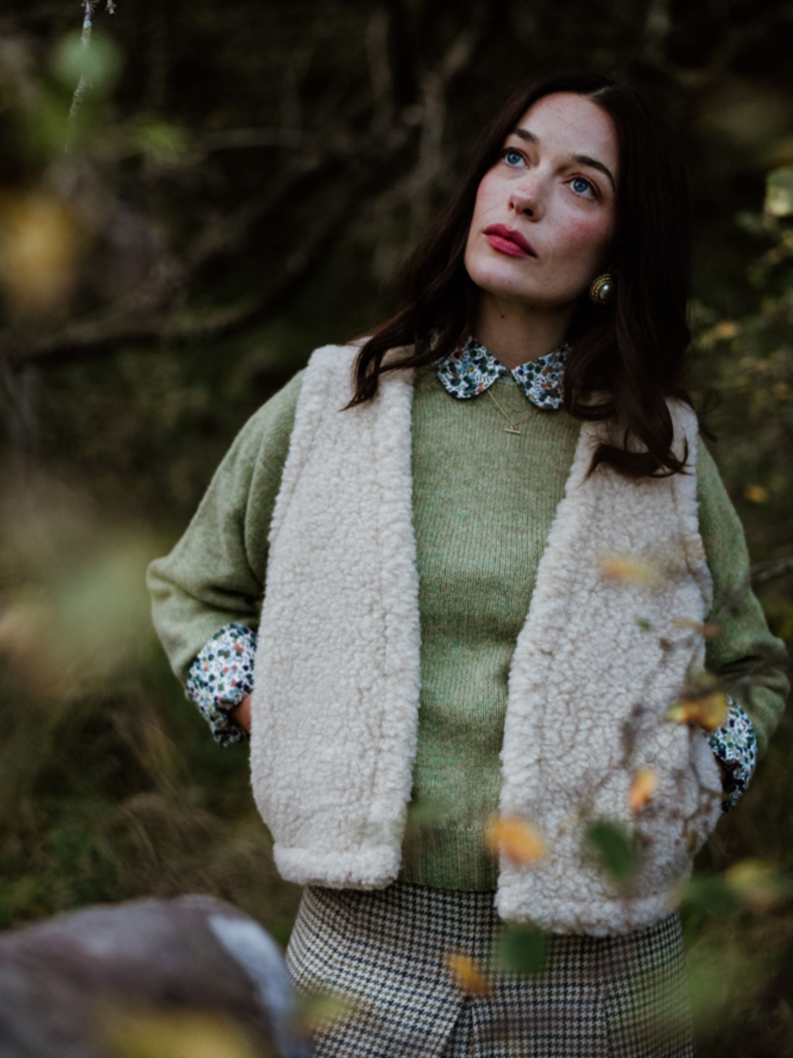 A woman with long brown hair stands outdoors, wearing the Campbell's of Beauly Wool Shrug over a green sweater and floral collared shirt. She gazes upward thoughtfully, surrounded by soft-focus greenery.