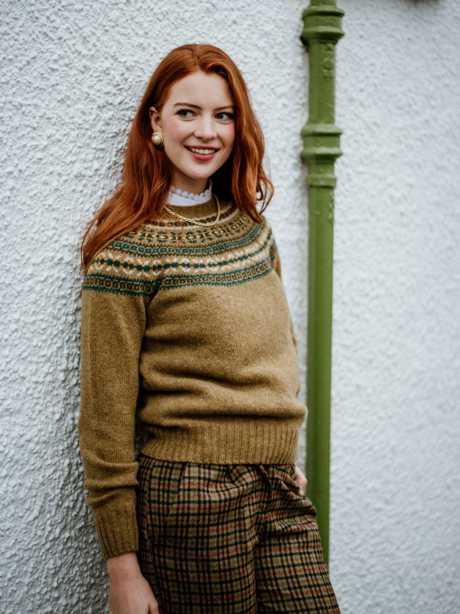 A smiling woman with long red hair wears the Campbells of Beauly Fairisle Crew Jumper in a traditional pattern and plaid pants, standing against a white textured wall with a green vertical pipe.