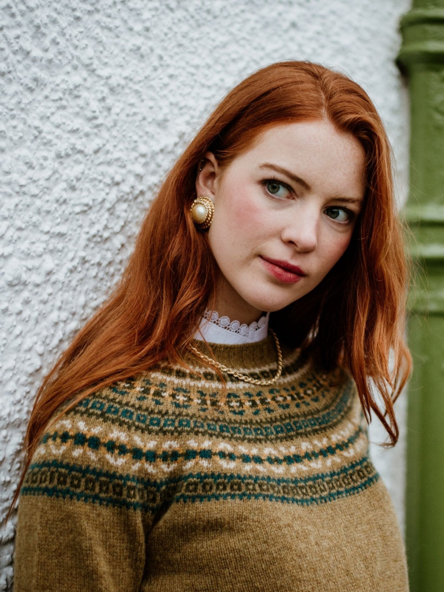 A young woman with long red hair stands against a textured white wall, wearing the Campbells of Beauly Fairisle Crew Jumper in a traditional pattern. She pairs it with vintage-style earrings and a white lace collar, gazing softly to the side.