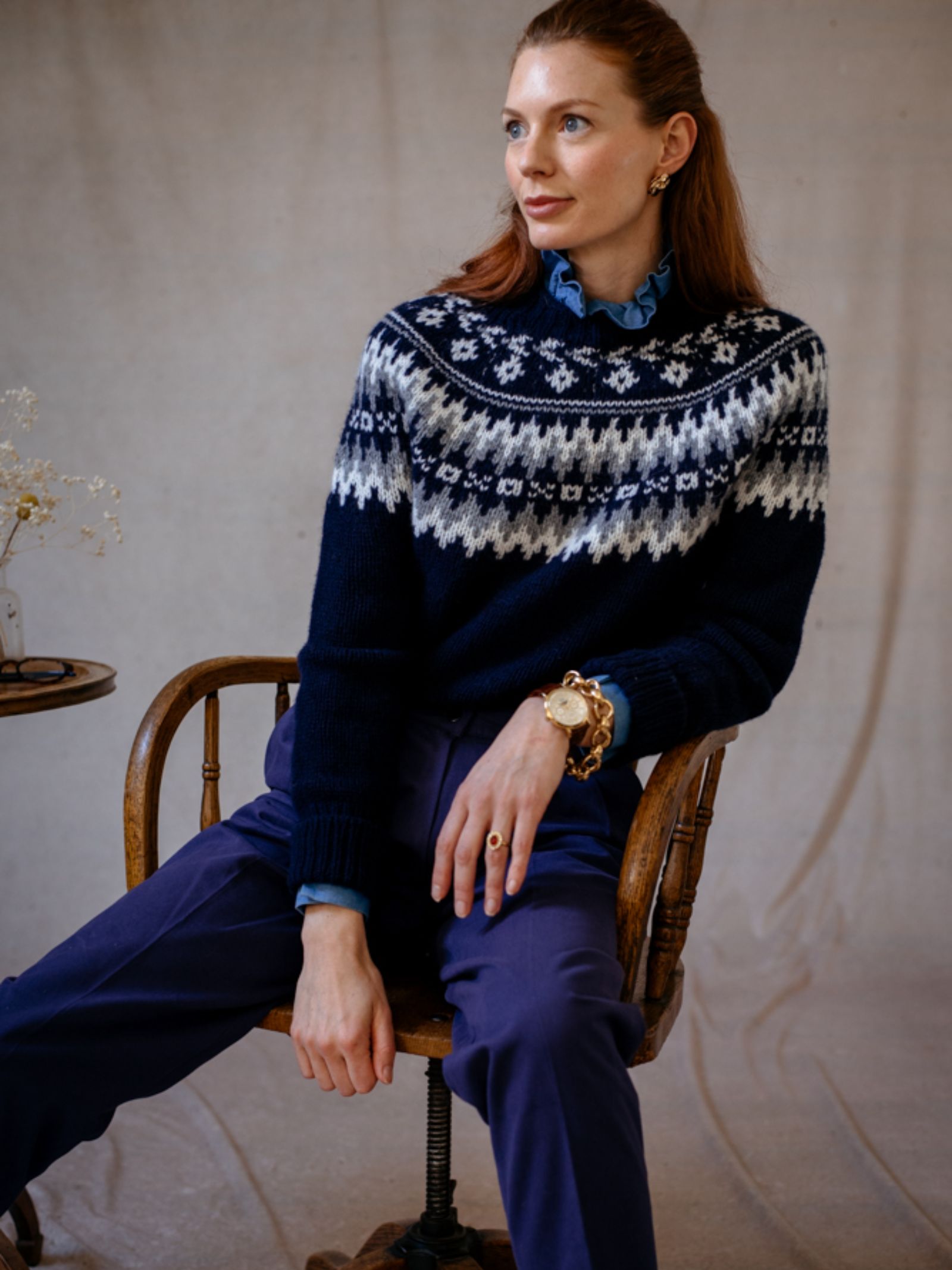 A woman with reddish-brown hair sits on a wooden chair, wearing Campbells of Beauly’s Chunky Fairisle Jumper in navy with a white Nordic pattern. She pairs it with blue trousers and a ruffled shirt, creating a cozy winter look in soft light.