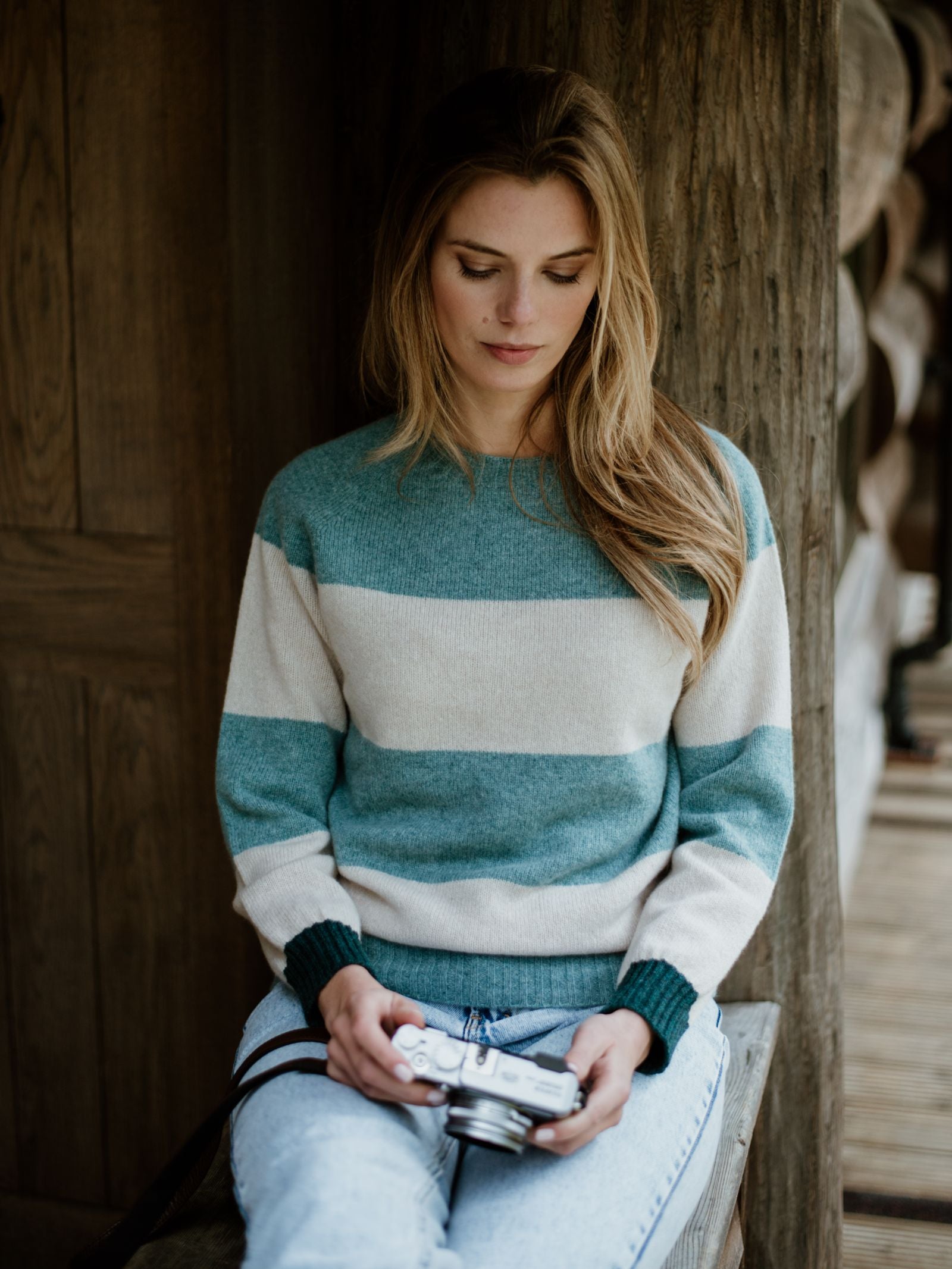 A woman with long blonde hair wears the Campbells of Beauly Stripe Crew Jumper, knitted in Scotland, and light blue jeans as she sits on a wooden bench holding a vintage camera, looking down calmly.