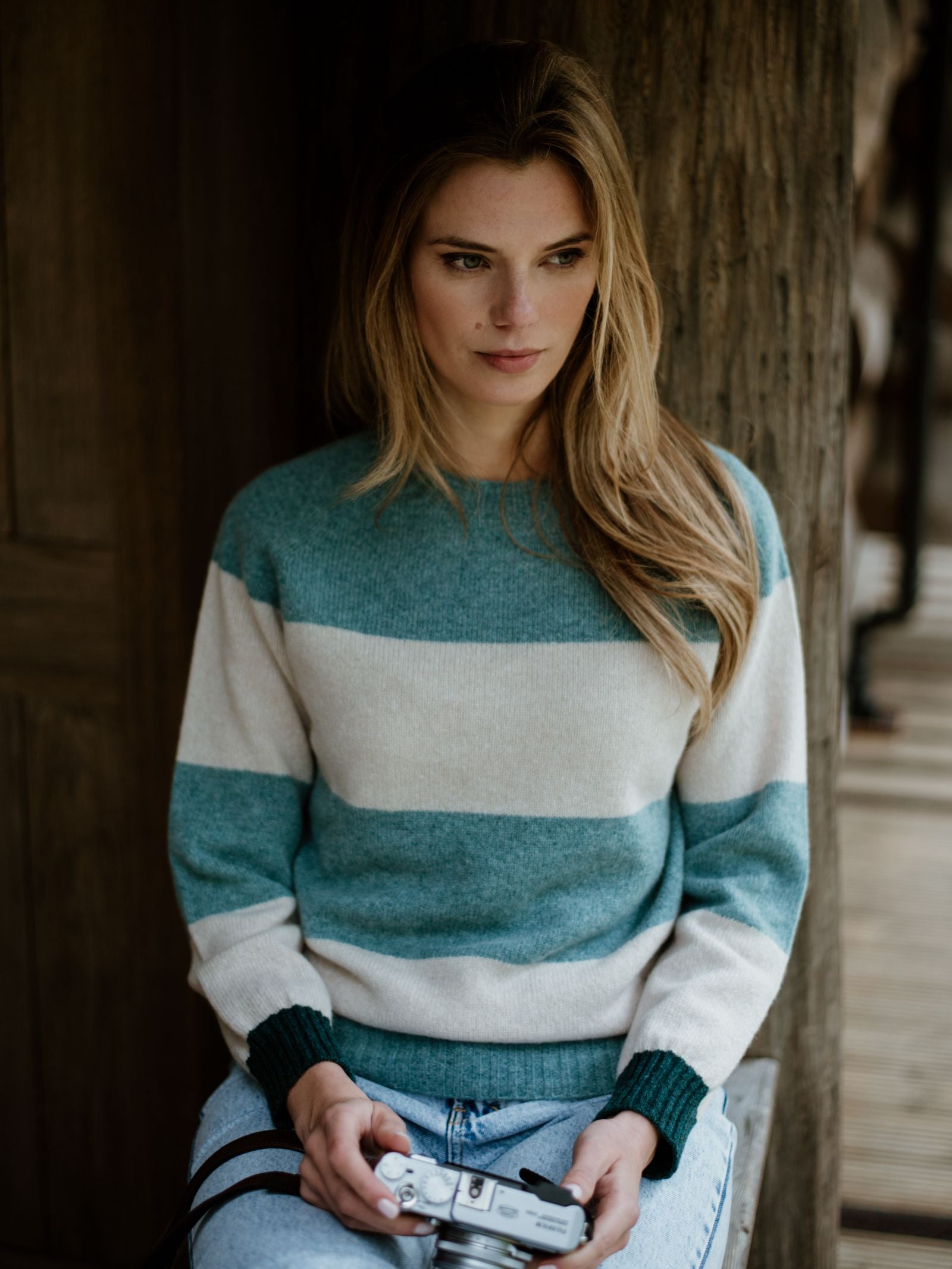 A woman with long, light brown hair wears the Stripe Crew Jumper by Campbells of Beauly, a blue and white knit crafted in Scotland. She sits indoors by a wooden wall, holding a vintage camera and looking thoughtfully to the side.