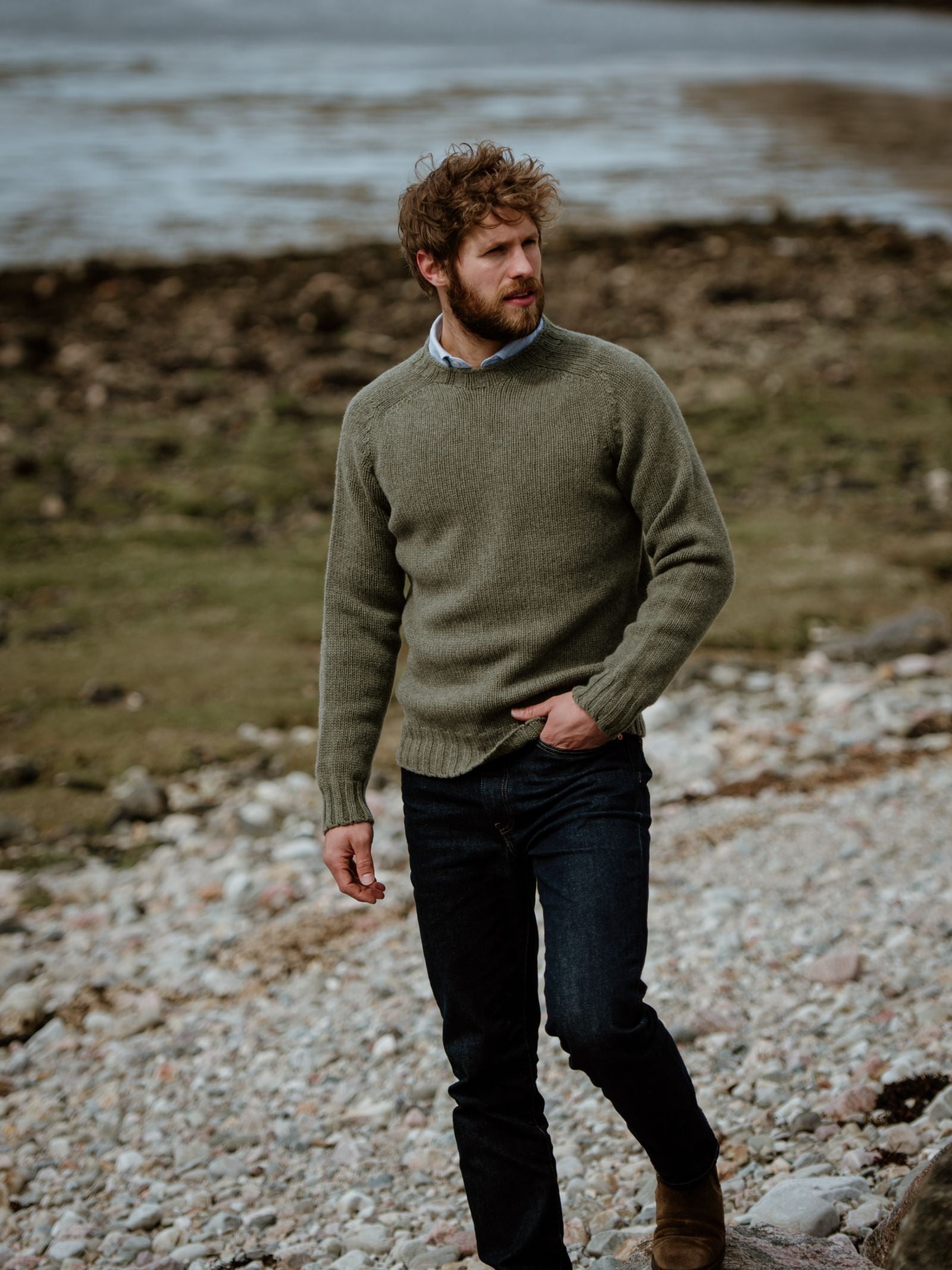 A man with curly hair and a beard walks on a rocky shoreline in blue jeans, brown boots, and a moss-green Campbells of Beauly Geelong Lambswool Chunky Crew Jumper. He looks to the side with one hand in his pocket against an overcast, blurred backdrop.