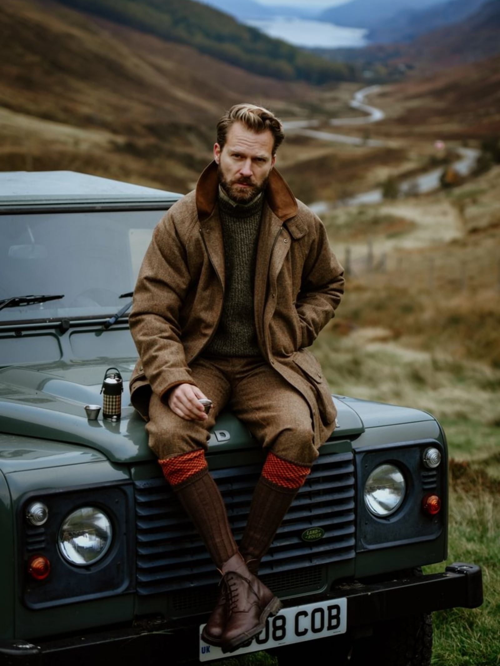 A man in brown outdoor gear sits on a green Land Rover with an 8oz Forest Green Hunter Flask by Campbell's of Beauly x Ettinger at his side, surrounded by rolling hills, a winding road, and a lake for a rustic atmosphere.