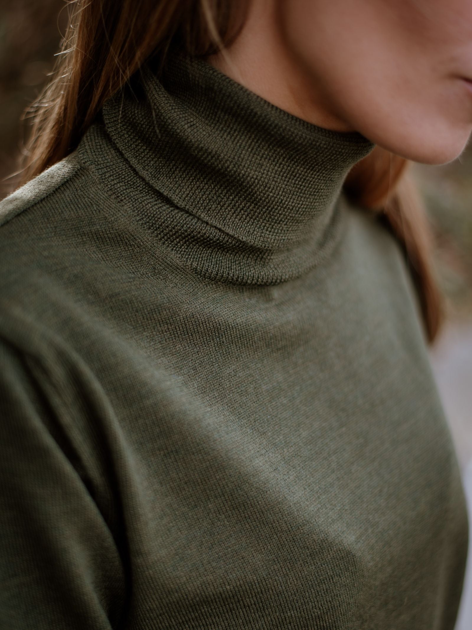 Close-up of a person wearing the Campbells of Beauly Merino Poloneck in olive green, focusing on the neck and shoulder to highlight the smart layering versatility of superfine merino wool, with the face blurred and partially out of frame.