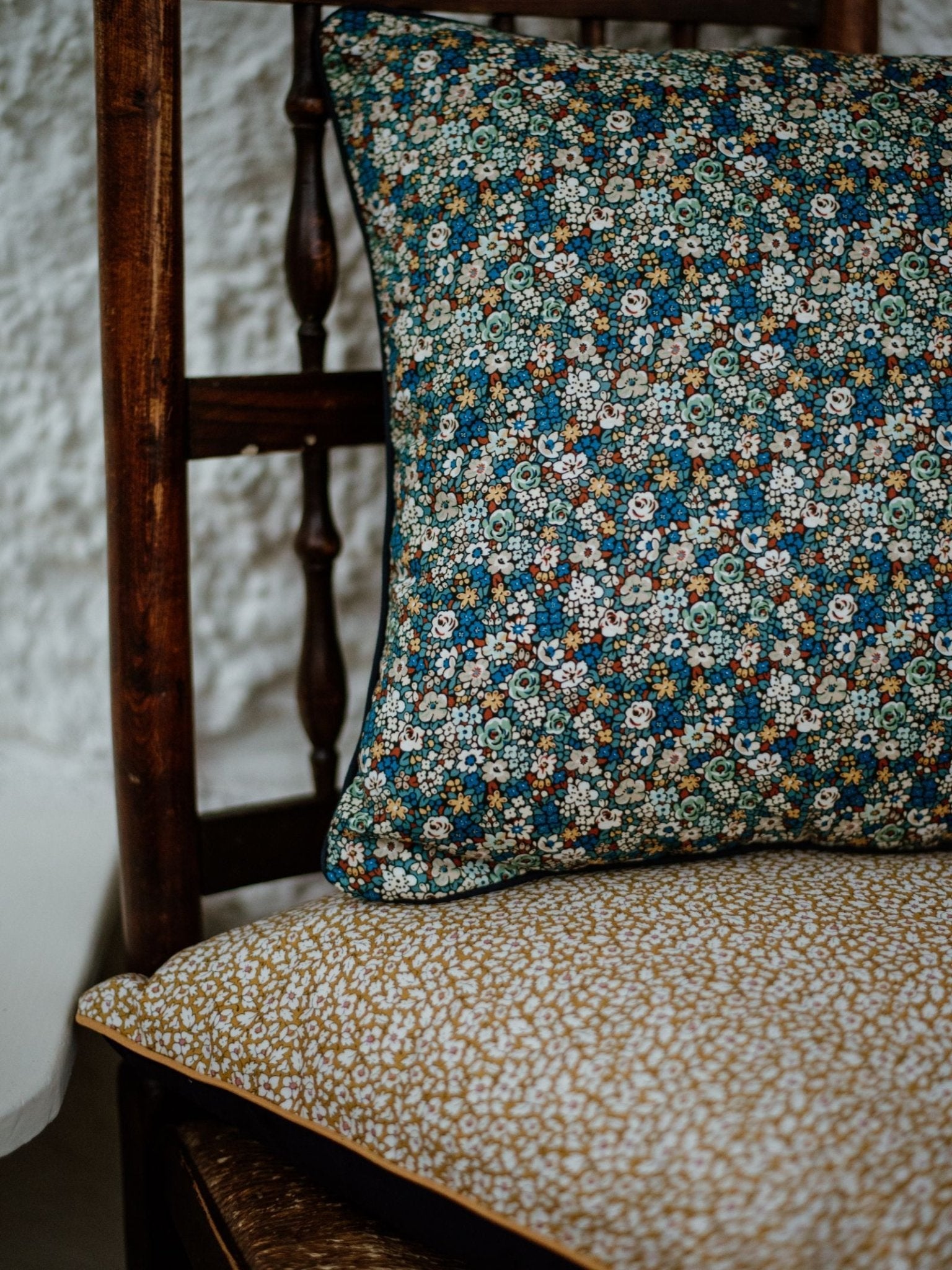 A wooden chair with a yellow floral seat cushion and a plush velvet blue Campbells of Beauly Liberty Print Cushion Cover, set against a textured white wall.