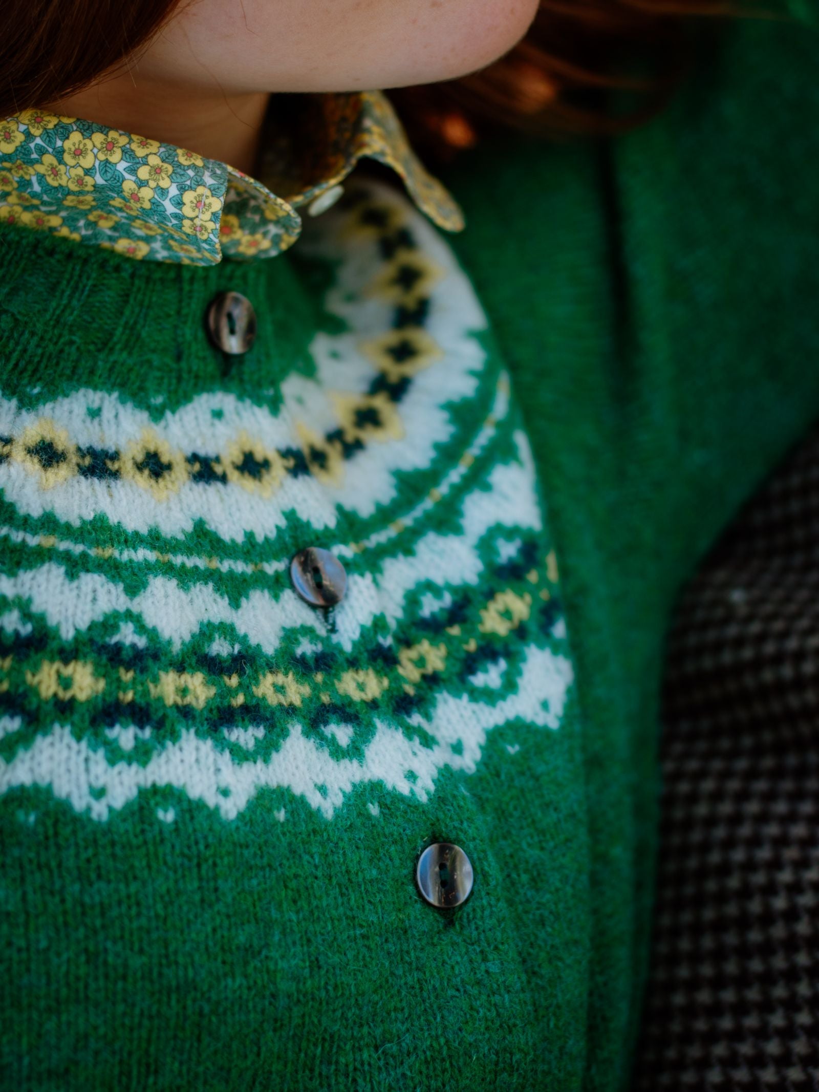 A person wears a green patterned Shetland Fairisle Cardigan by Campbells of Beauly with black buttons and a floral collared shirt underneath. Only their lower face and upper torso are visible, highlighting classic knitwear style.