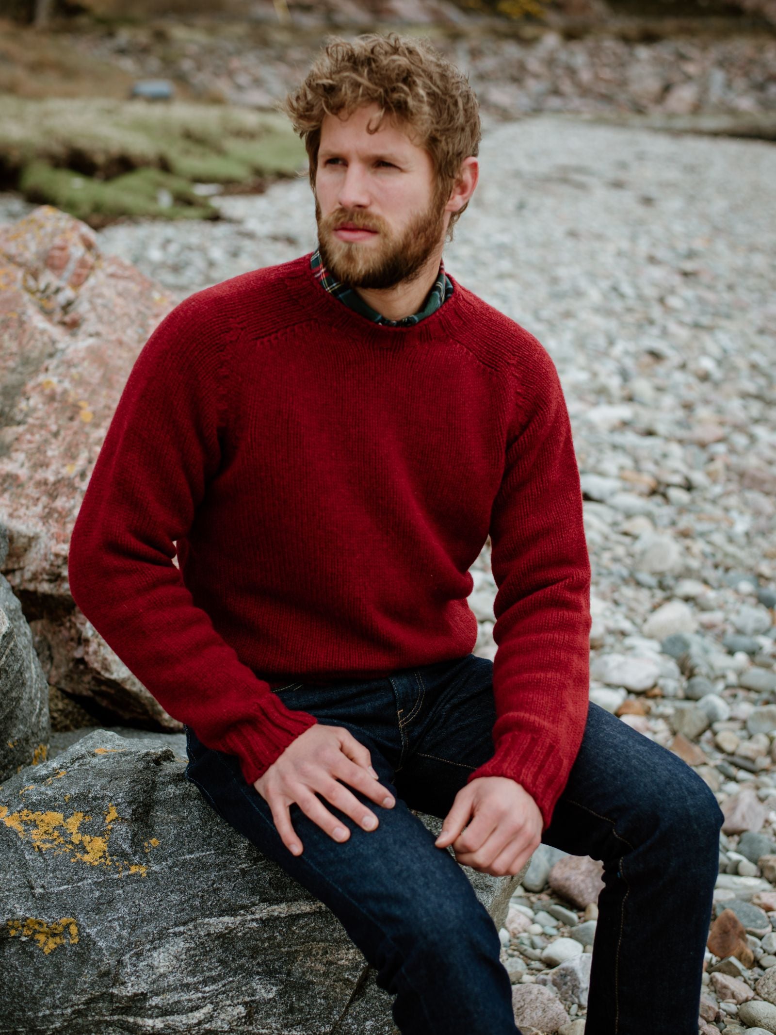 A bearded man with curly hair wears the Campbells of Beauly Geelong Lambswool Chunky Crew Jumper over a plaid shirt and dark jeans, sitting thoughtfully on a large rock in a natural, rocky outdoor landscape.