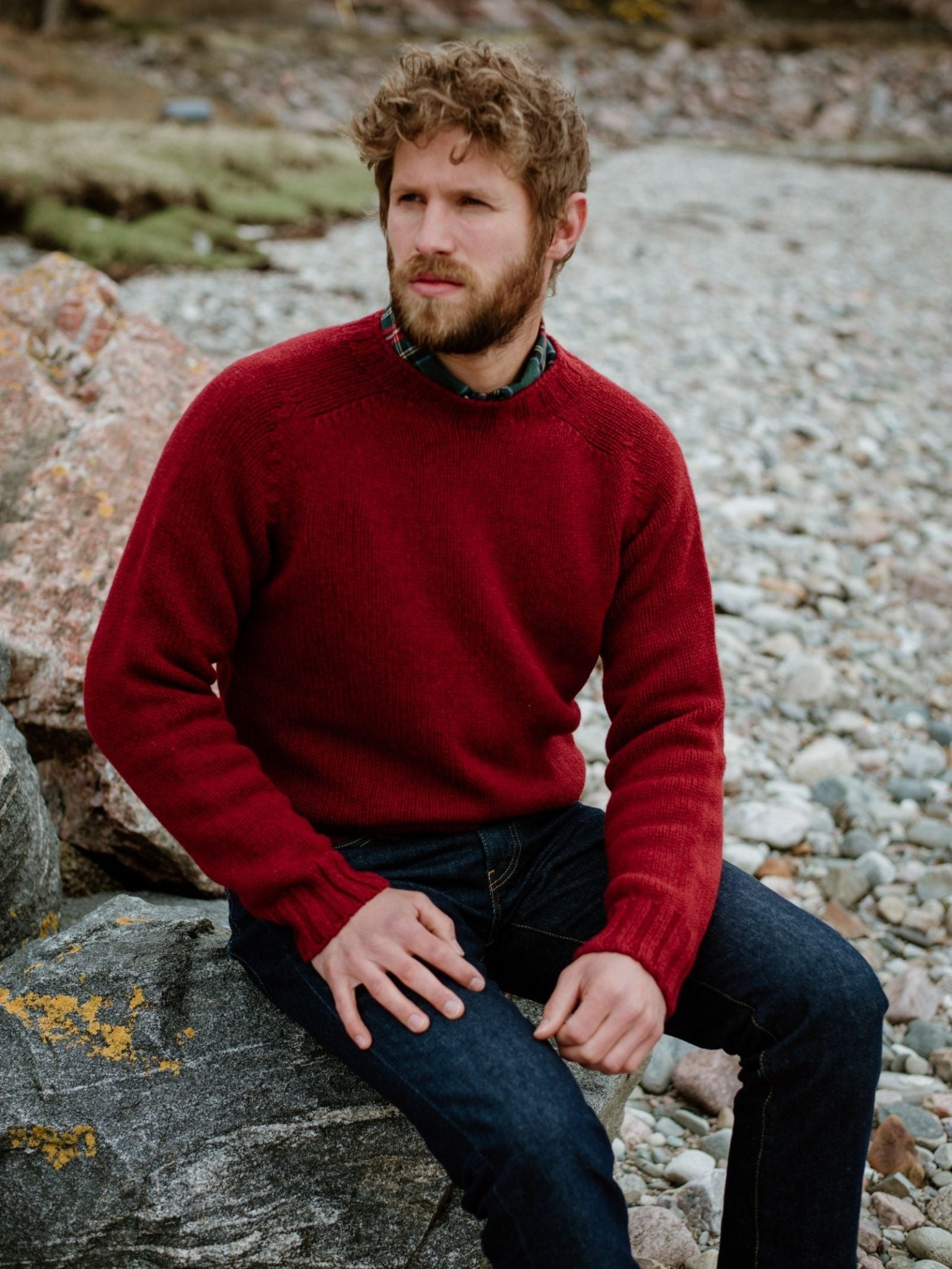 A man with curly hair and a beard wears the Campbells of Beauly Geelong Lambswool Chunky Crew Jumper in red and dark jeans, sitting thoughtfully on a large rock by a rocky shoreline.