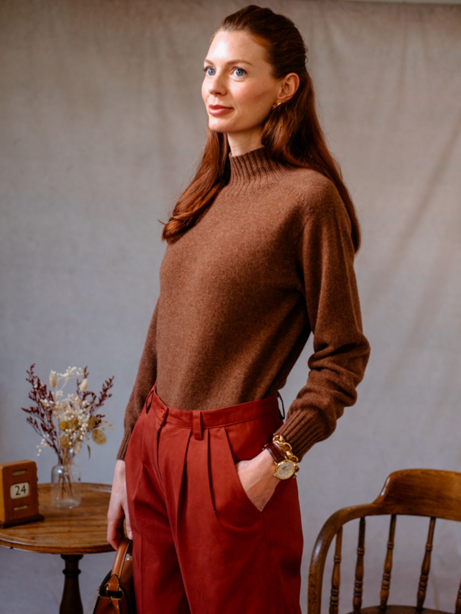 A woman with long red hair stands indoors, wearing the Campbell's of Beauly Auld Stock Geelong Lambswool Funnel Neck Jumper and rust-colored pants. One hand is in her pocket as she stands beside a wooden table with flowers and a wooden chair.