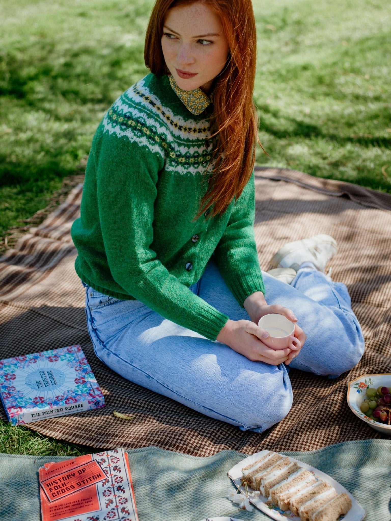 A person with long red hair wearing the Campbells of Beauly Shetland Fairisle Cardigan and light blue jeans sits on a blanket outdoors, holding a cup, with books, grapes, and cake slices nearby.