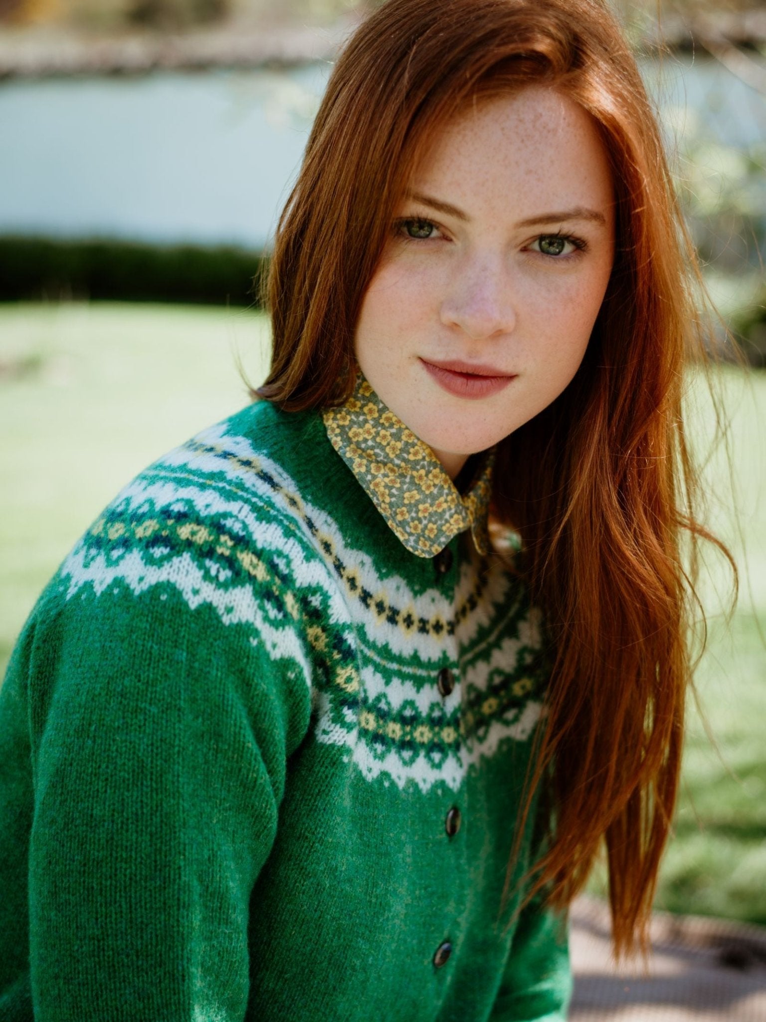 A young woman with long red hair, light skin, and freckles wears a Campbells of Beauly Shetland Fairisle Cardigan in pure new wool over a yellow floral collared shirt, sitting outdoors on a sunny day.
