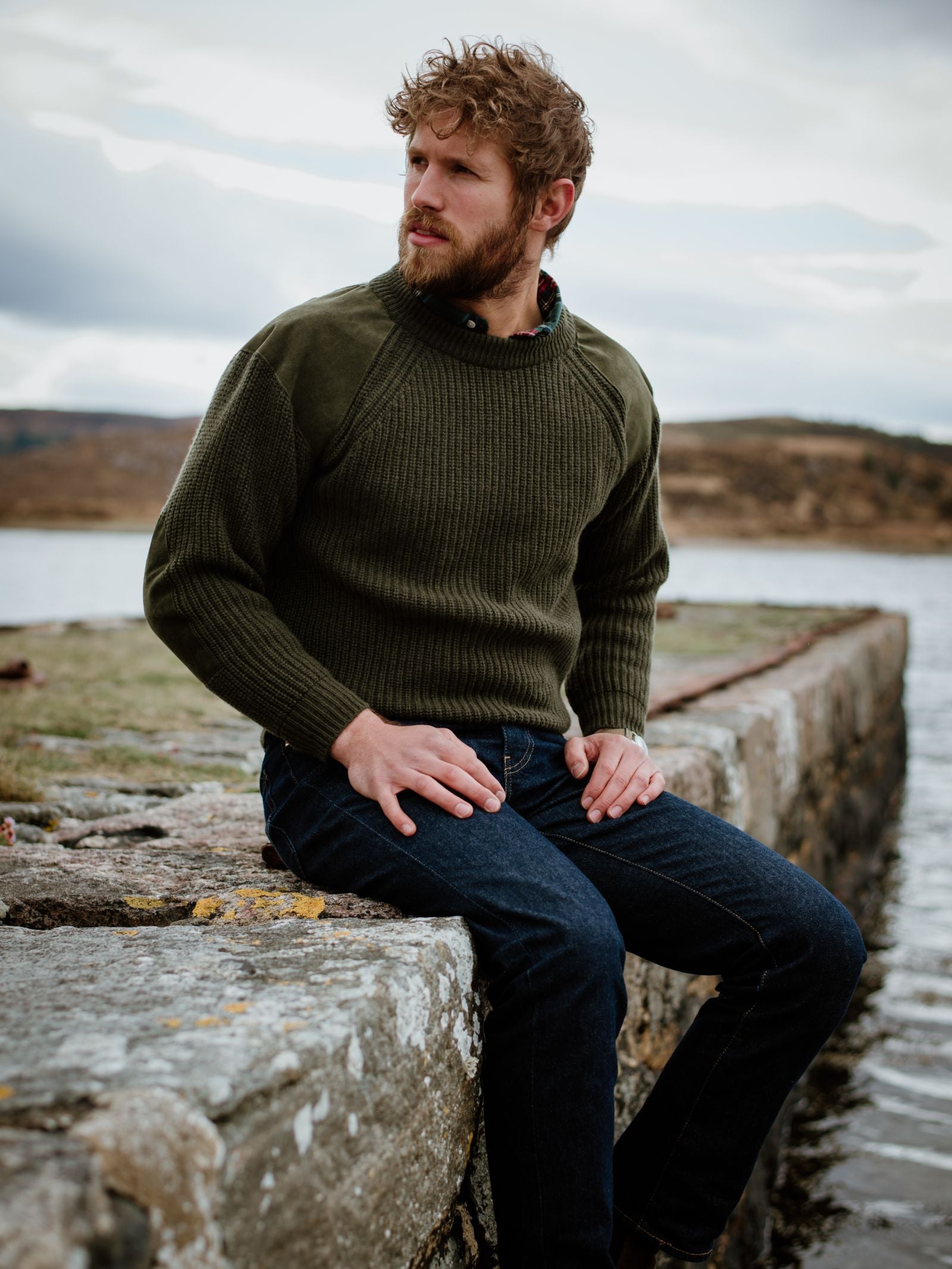 A bearded man with curly hair wears the Lux Cashmere Shooter Crew Jumper by Campbells of Beauly and dark jeans, sitting on a stone wall by the water against a natural landscape and cloudy sky, showcasing timeless country style.