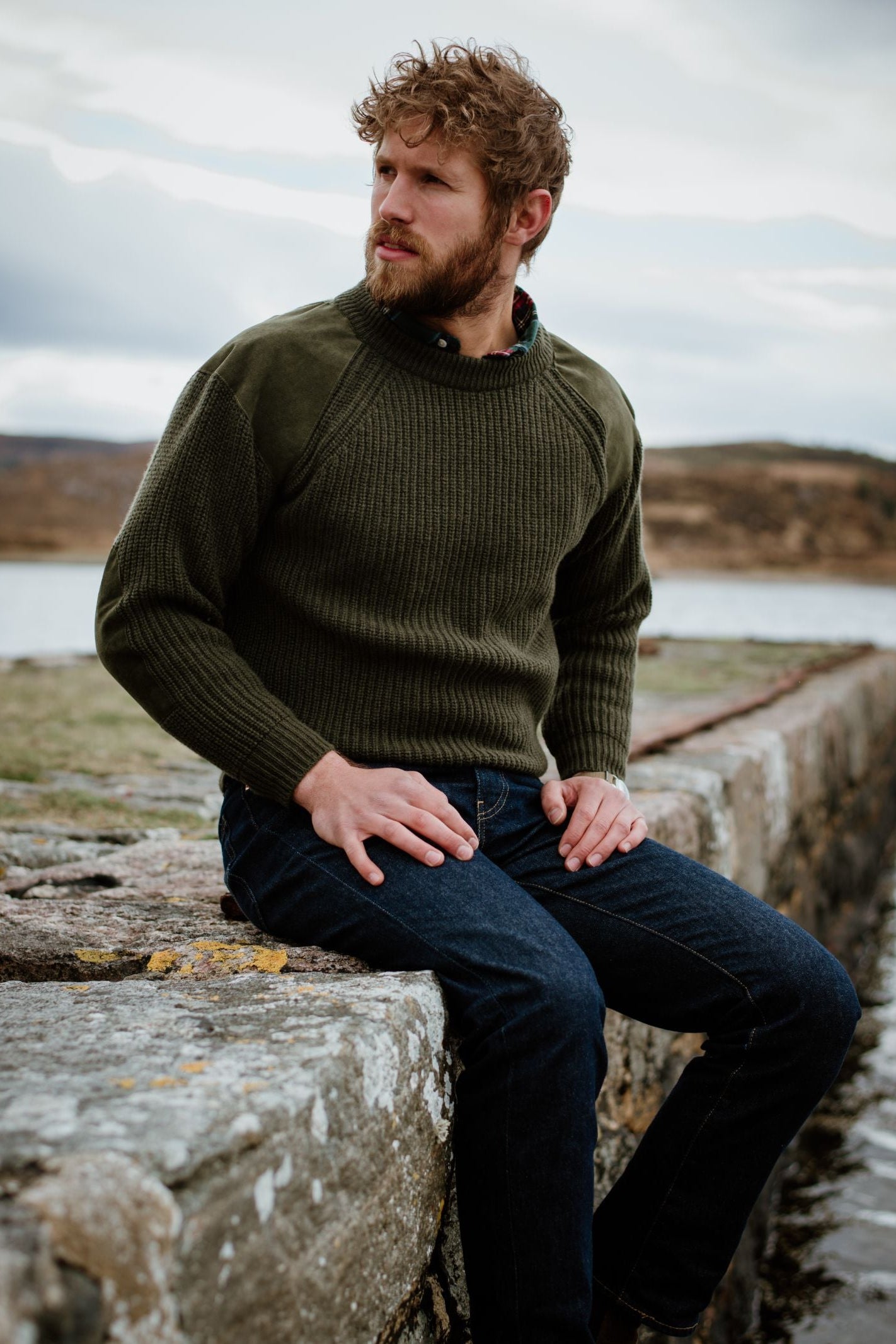 A bearded man with curly hair wears the Lux Cashmere Shooter Crew Jumper by Campbells of Beauly and dark jeans, sitting on a stone wall by the water against a natural landscape and cloudy sky, showcasing timeless country style.