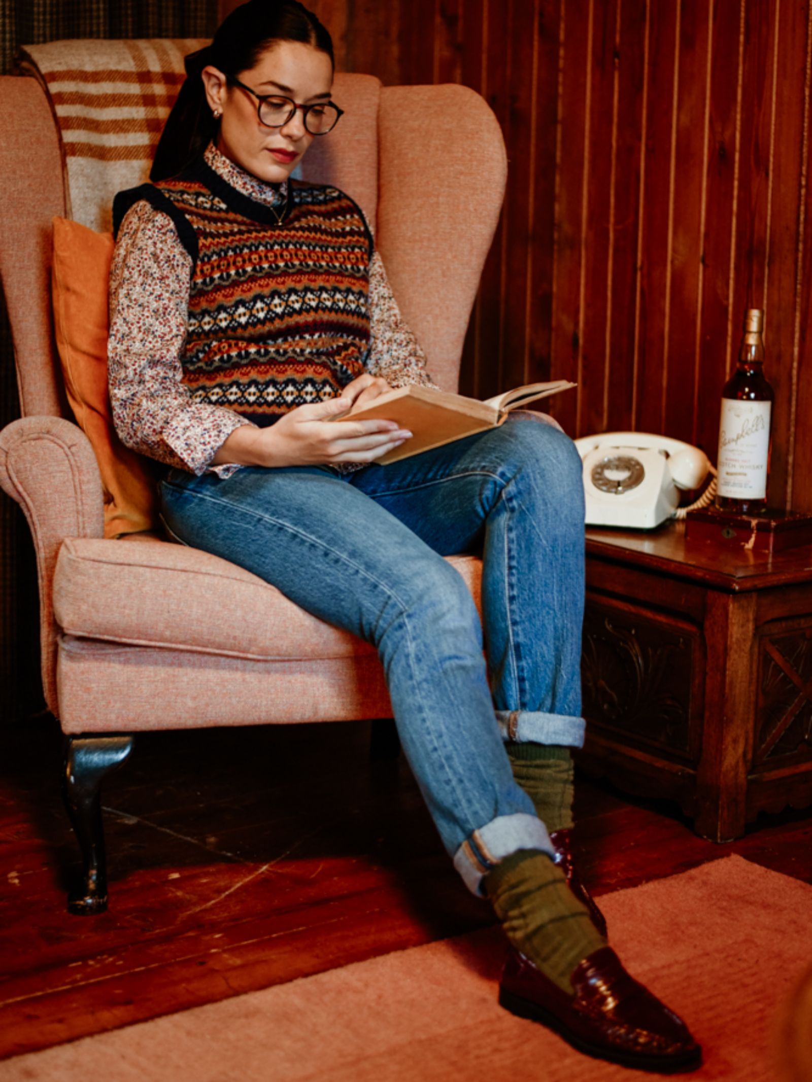 A woman in Campbell's of Beauly Rib Cashmere Socks, glasses, a patterned sweater vest, and jeans reads in a pink armchair. A vintage rotary phone and liquor bottle rest on a wooden table beside her, creating a cozy retro scene.