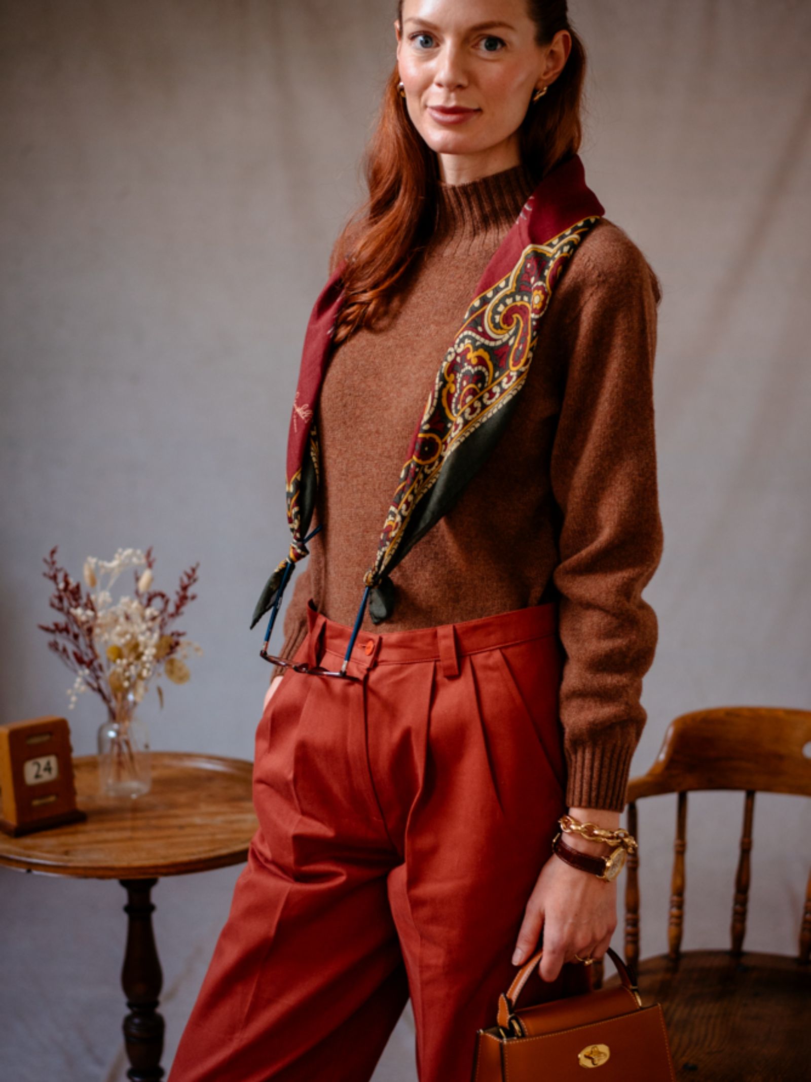A woman with long red hair wears a brown sweater, rust trousers, and the Campbells of Beauly Wool & Silk Scarf. She holds a small brown handbag and stands beside a round table with dried flowers and a wooden chair.