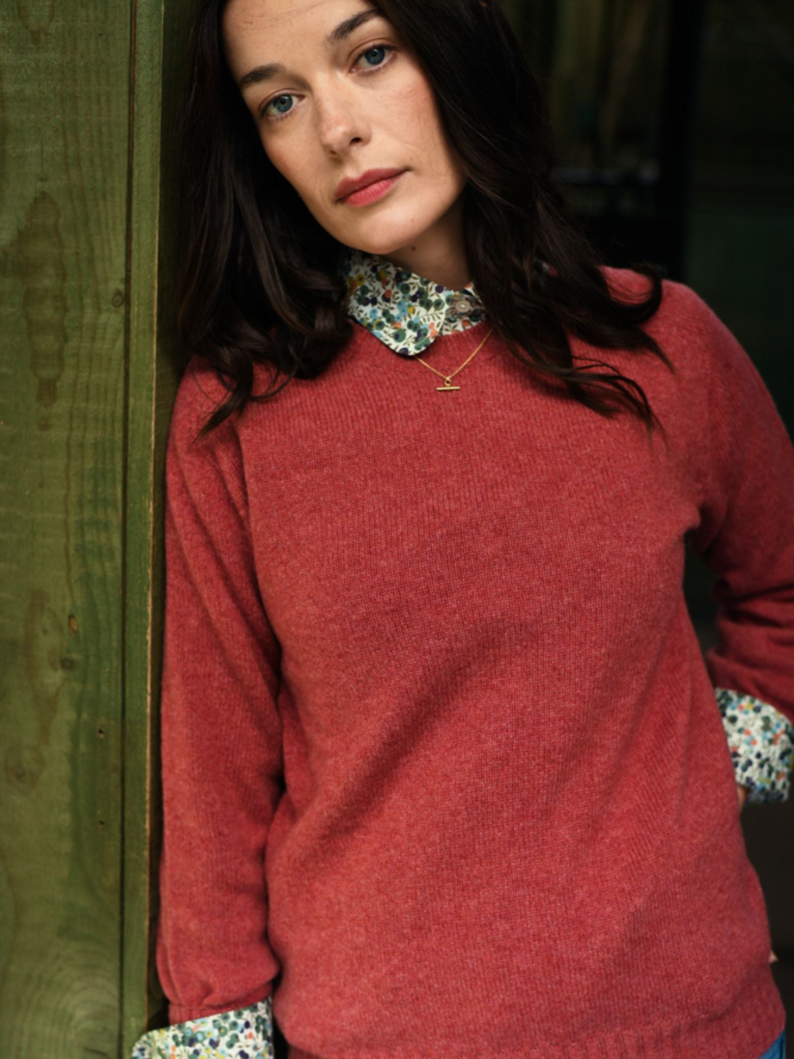 A woman with long dark hair leans against a green wooden wall, wearing the Campbell's of Beauly Geelong Lambswool Crew Neck Jumper over a floral collared shirt and delicate gold necklace, gazing softly at the camera with a relaxed expression.