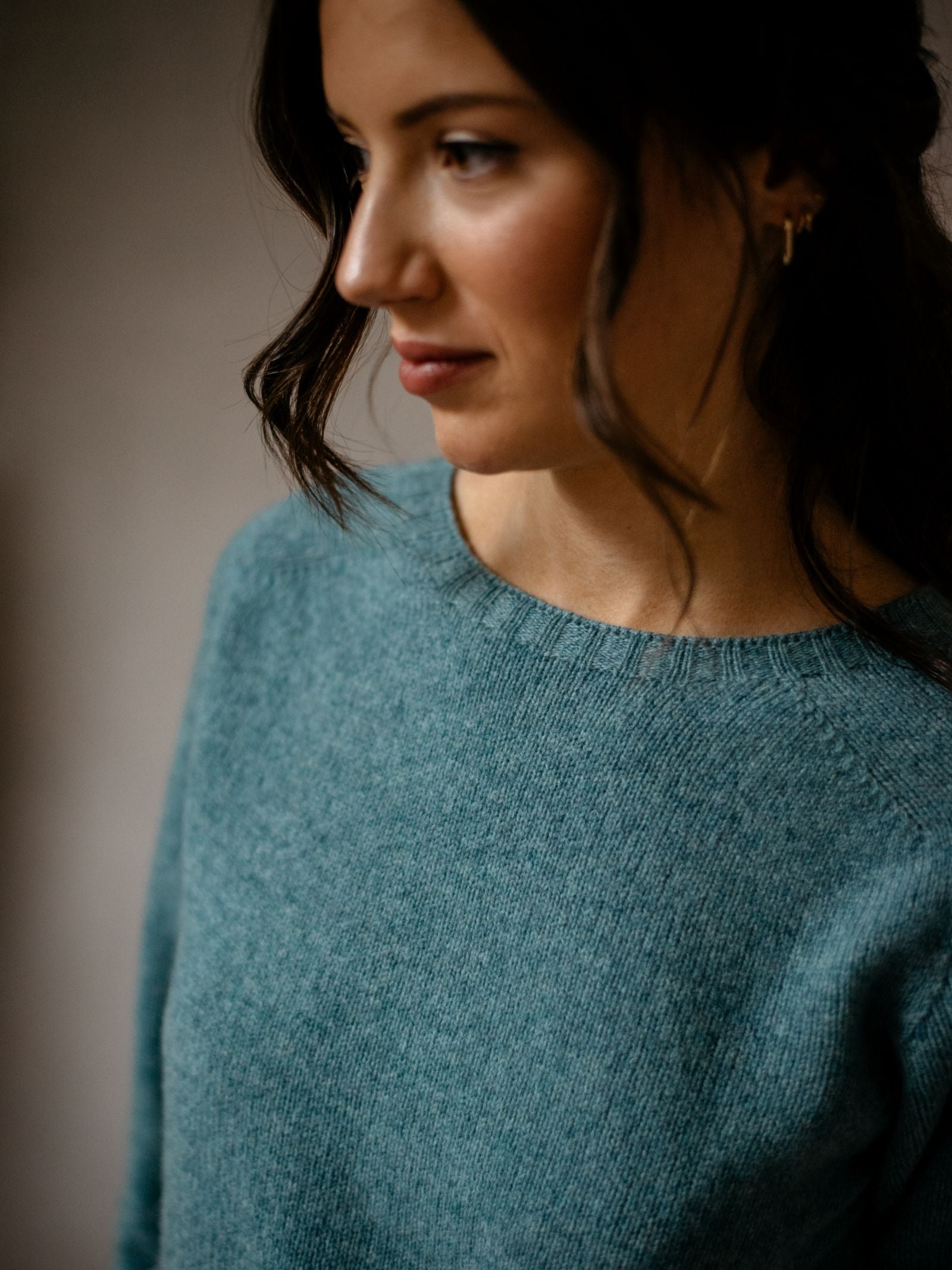 A woman with wavy dark hair wears a teal Geelong Lambswool Crew Neck Jumper by Campbells of Beauly, gazing to the side with a contemplative expression against a softly blurred neutral background.