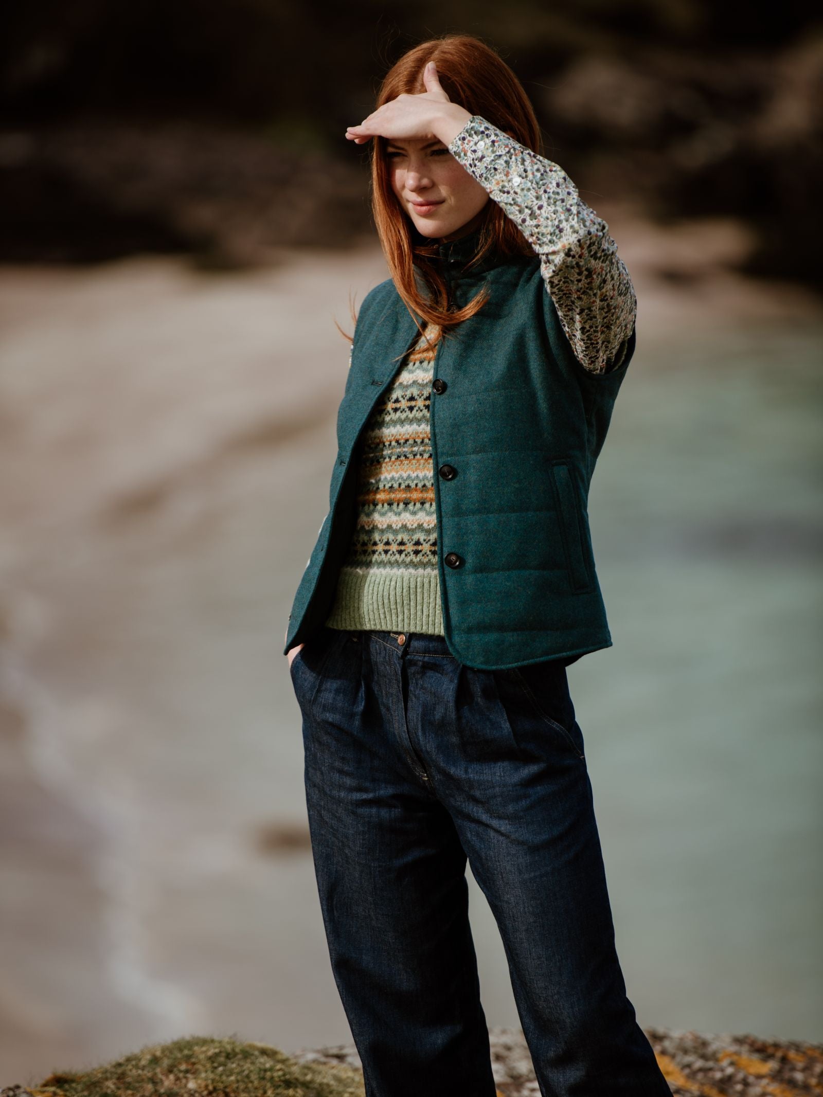 A woman with long red hair stands near the beach, wearing the Campbells of Beauly Quilted Buttoned Gilet over a patterned sweater and dark jeans. She shields her eyes from the sun with one hand, gazing into the distance.