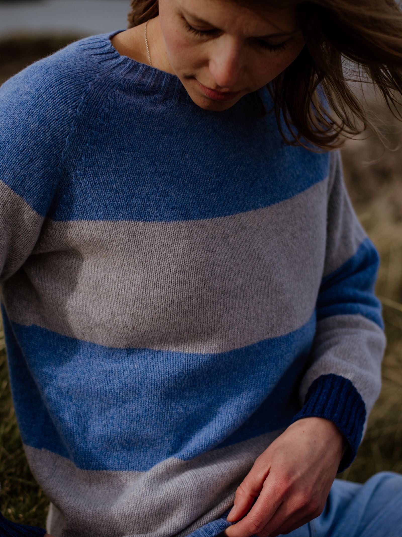 A person with light brown hair wears the Stripe Crew Jumper by Campbells of Beauly—a blue and gray striped crew neck, expertly knitted in Scotland from quality yarns—while sitting outdoors in natural light.