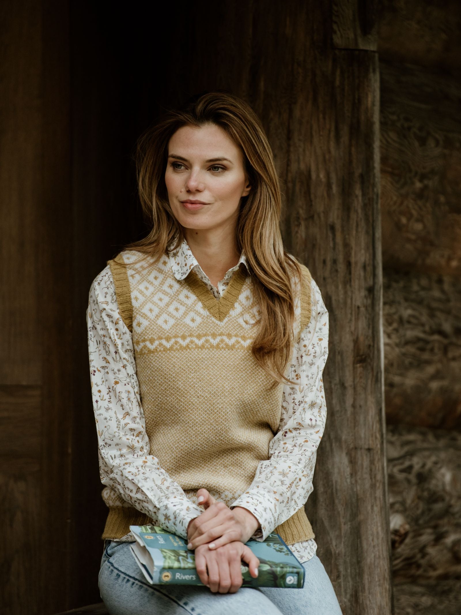 A woman with long brown hair, wearing the Campbells of Beauly Nordic V Tank over a floral shirt and jeans, sits outdoors against a wooden wall, holding a book in her lap and gazing thoughtfully to the side.