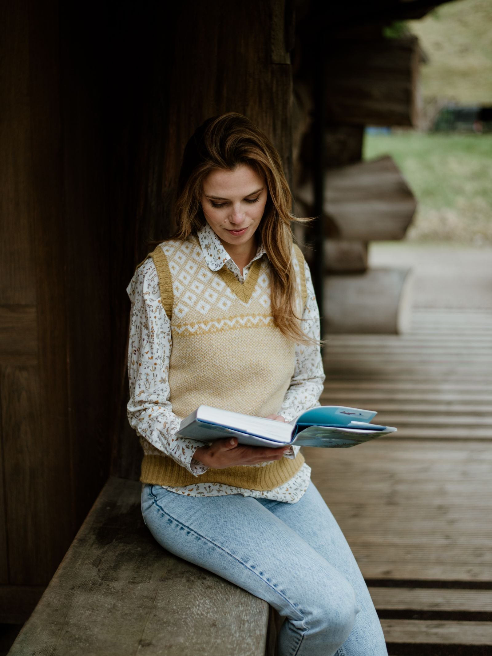 A woman with light brown hair, wearing the Campbells of Beauly Nordic V Tank and light blue jeans, sits on a wooden bench outdoors reading a book, with a blurred wooden structure in the background.