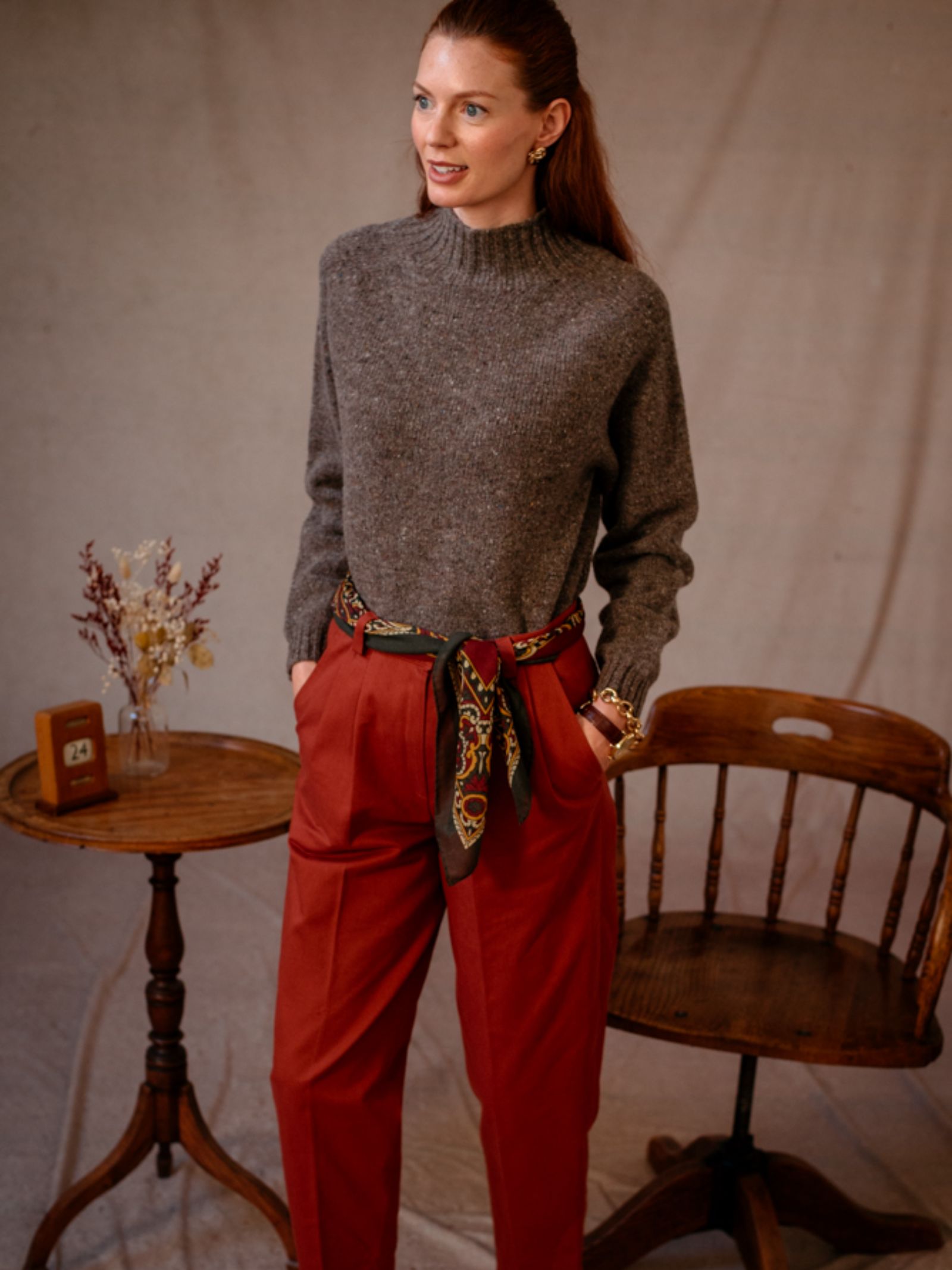 A woman stands indoors wearing Campbells of Beauly Donegal Lambswool Funnel Neck jumper with flecks, high-waisted red trousers, and a patterned scarf as a belt, near a wooden chair and round table with dried flowers and a small box.