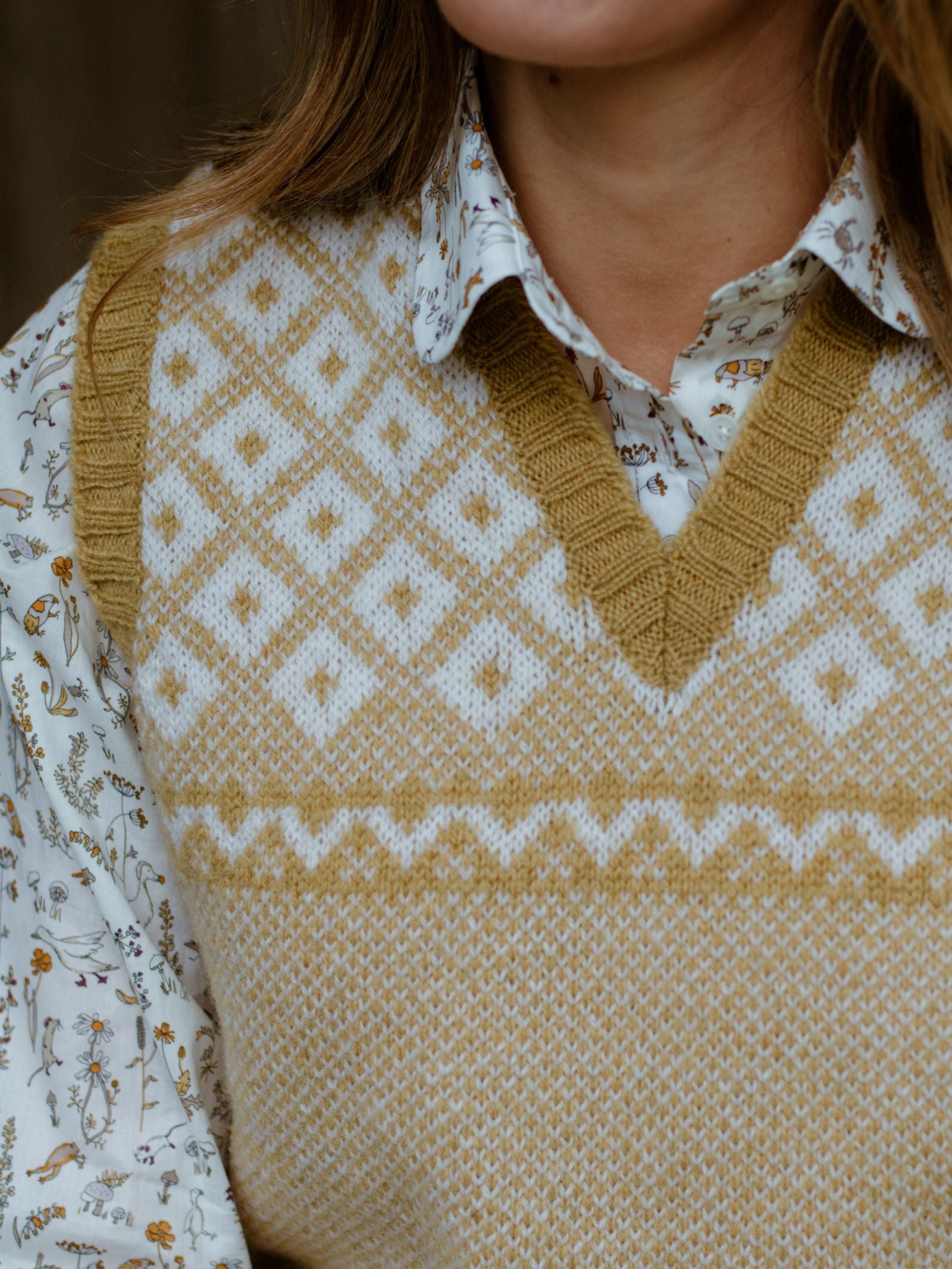 A close-up of a person in a white botanical print shirt layered with the Campbells of Beauly Nordic V Tank, a V-neck sweater vest featuring tan and white Fair Isle diamond and zigzag patterns; the face is partially visible.