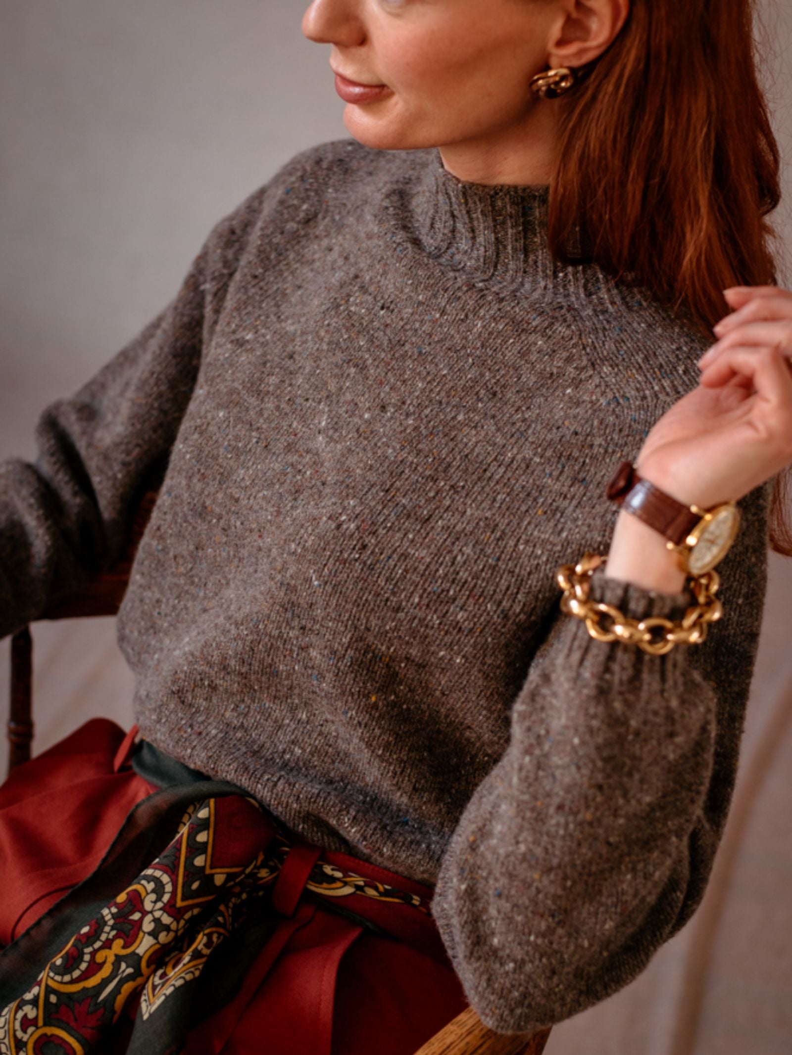 A woman with long reddish hair wears the Campbells of Beauly Donegal Lambswool Funnel Neck jumper, paired with a patterned scarf, rust skirt, gold earrings, chunky gold bracelet, watch, and ring.
