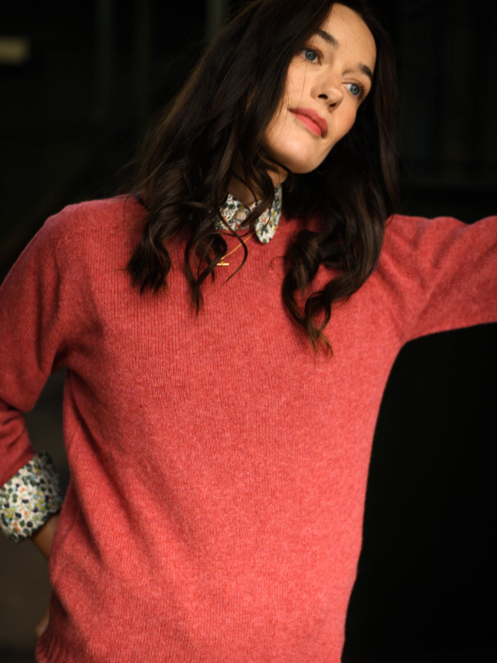 A woman with long, wavy dark hair wears the Campbell's of Beauly Geelong Lambswool Crew Neck Jumper in pink over a floral collared shirt, standing with one arm raised against a dark, blurred background.