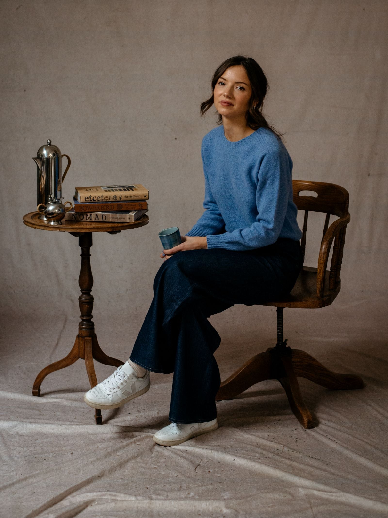 A woman in a blue Campbells of Beauly Geelong Lambswool Crew Neck Jumper and dark pants sits on a wooden chair, holding a blue cup. Next to her is a round table with books and coffee items, set against a neutral backdrop. Made in Scotland.