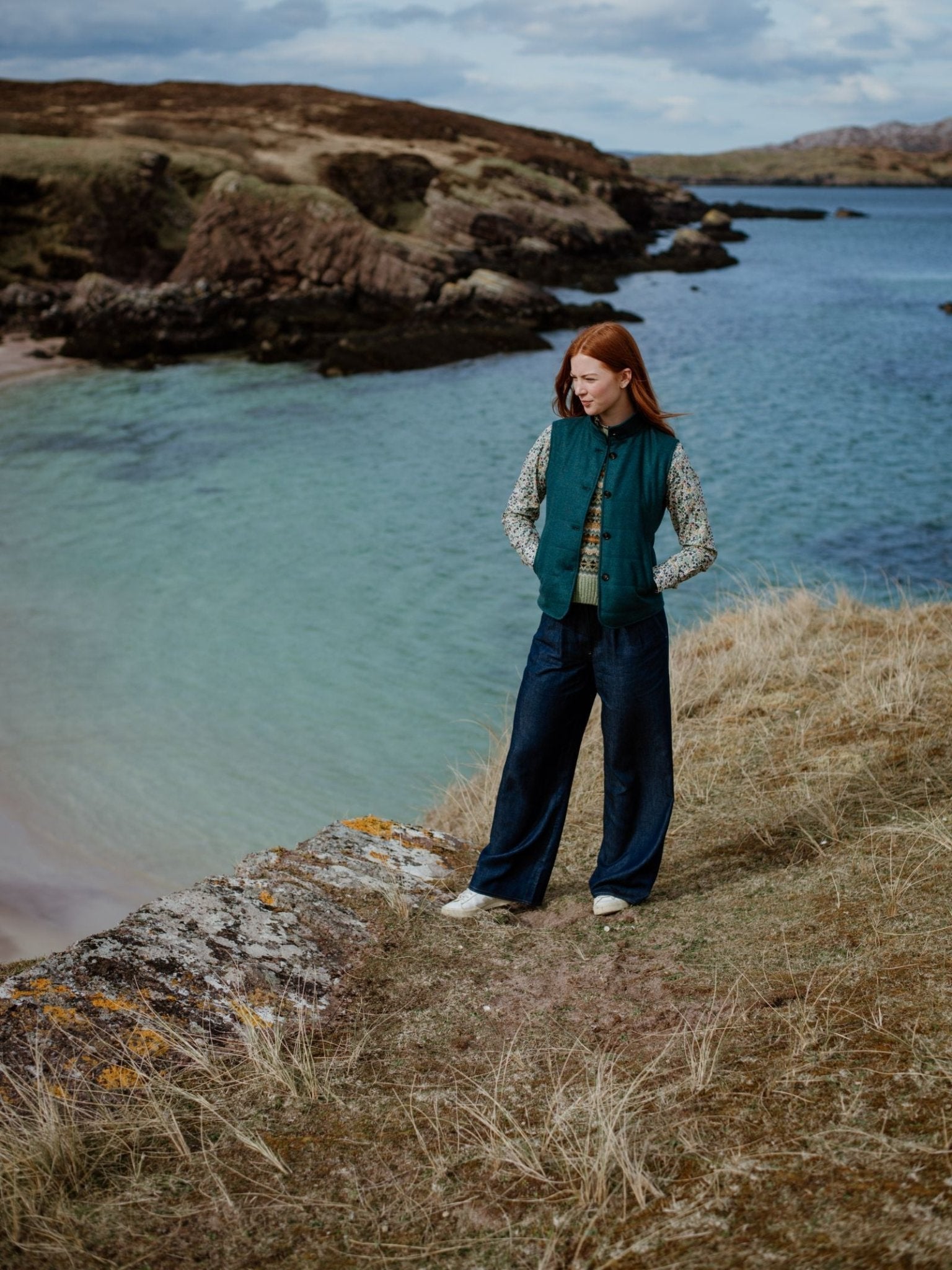 A person with long red hair stands on a grassy hillside by clear blue water, wearing the Campbells of Beauly Quilted Buttoned Gilet over a patterned shirt and wide-leg jeans—a versatile layering piece for rugged landscapes.