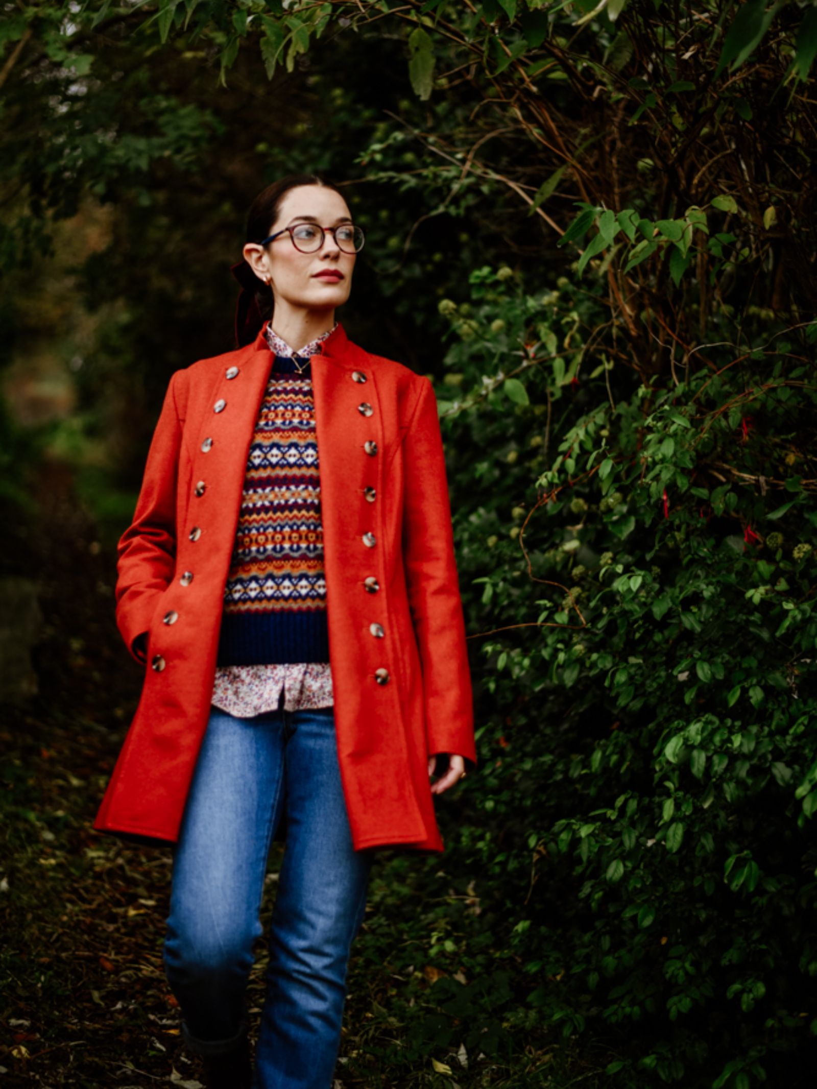 A person wearing glasses, a Campbell's of Beauly Pirate Coat in red, a colorful patterned sweater, and blue jeans stands outdoors on a leafy path beside dense green foliage, looking to the side.