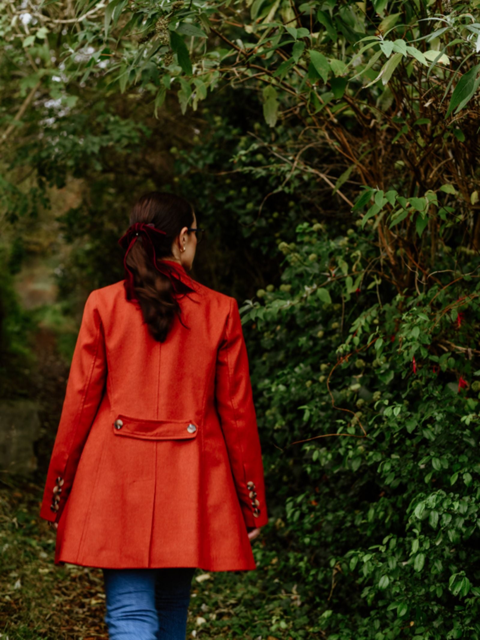 A person with long hair tied back, wearing a bright red Campbell's of Beauly Pirate Coat and blue jeans, walks through a green outdoor area filled with bushes and trees, facing away from the camera.