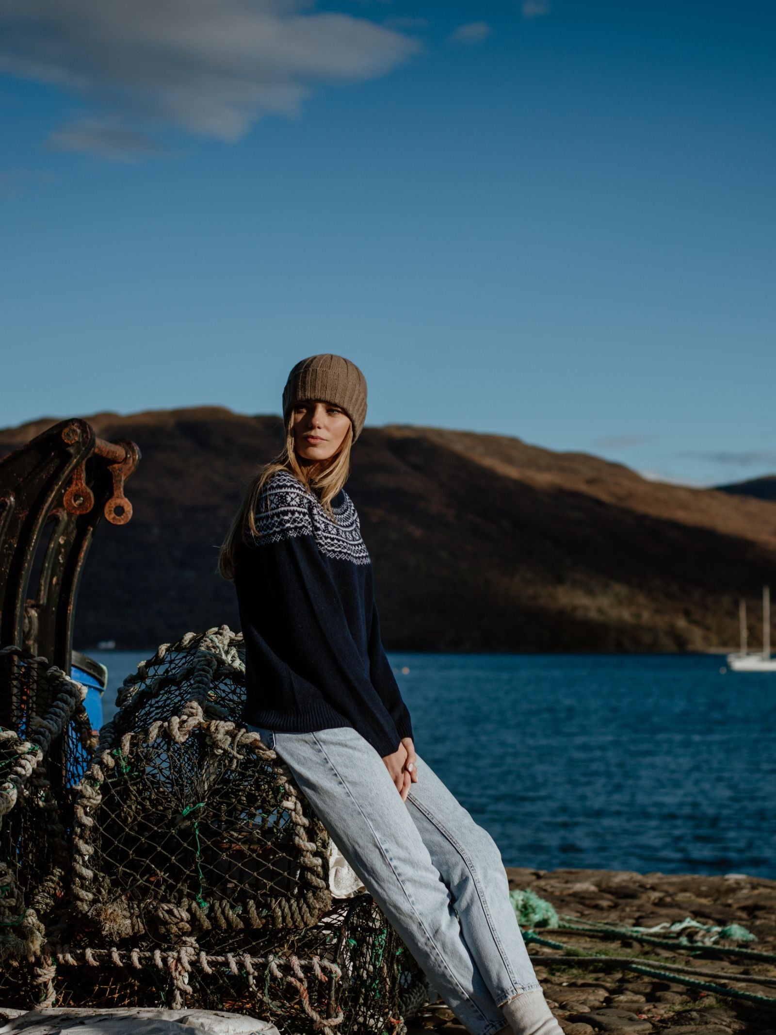 A woman wearing Campbells of Beaulys Two-Colour Fairisle Yoke Jumper and a knit hat sits on fishing nets by the water, with hills and a sailboat in the background under a blue sky.