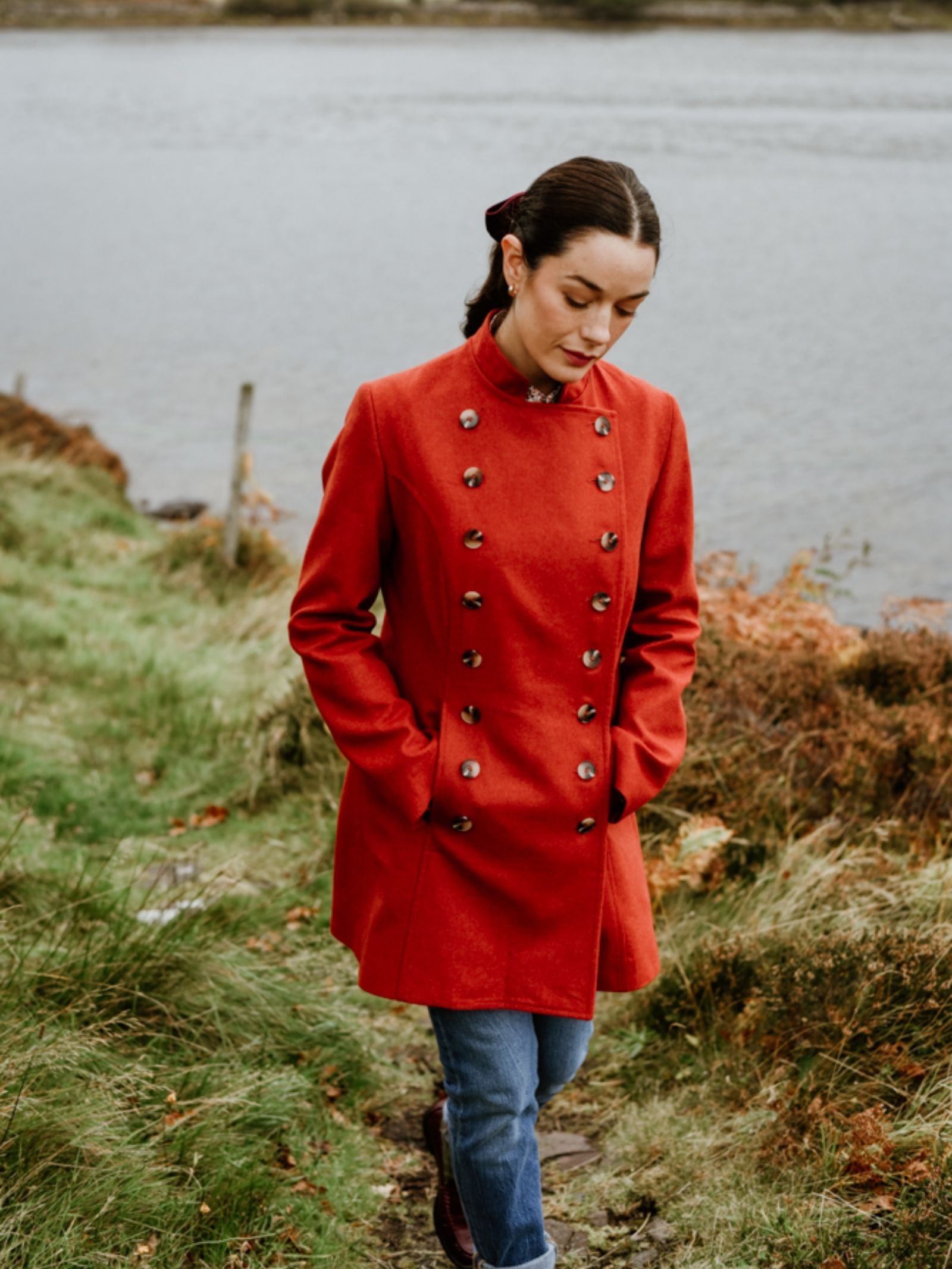 A woman in a bold red Campbell's of Beauly Pirate Coat and blue jeans walks along a grassy path by the water, hands in pockets and head slightly bowed.