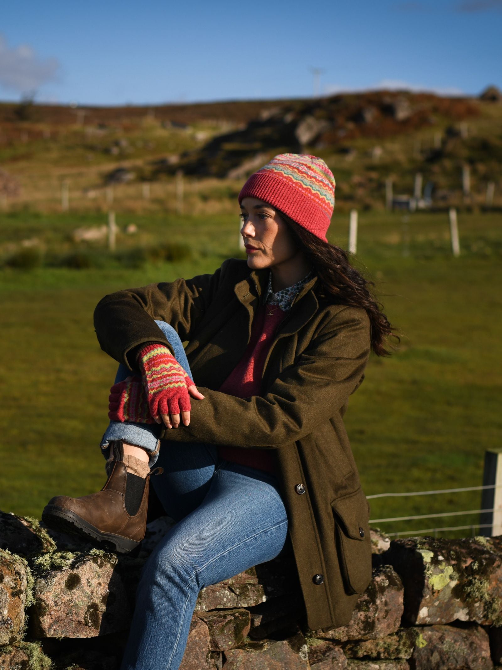 A woman wearing Campbell's of Beauly Lambswool Fairisle Half-Finger Gloves, a striped Scottish knit hat, olive coat, blue jeans, and brown boots sits on a stone wall in a rural landscape with grassy hills behind her.